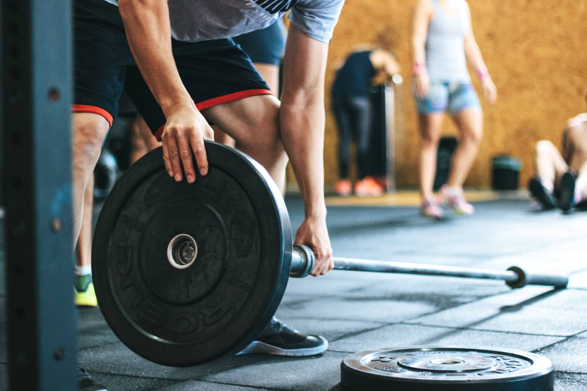 Person in gym attire placing a weight plate on a barbell, with other people visible in the background.