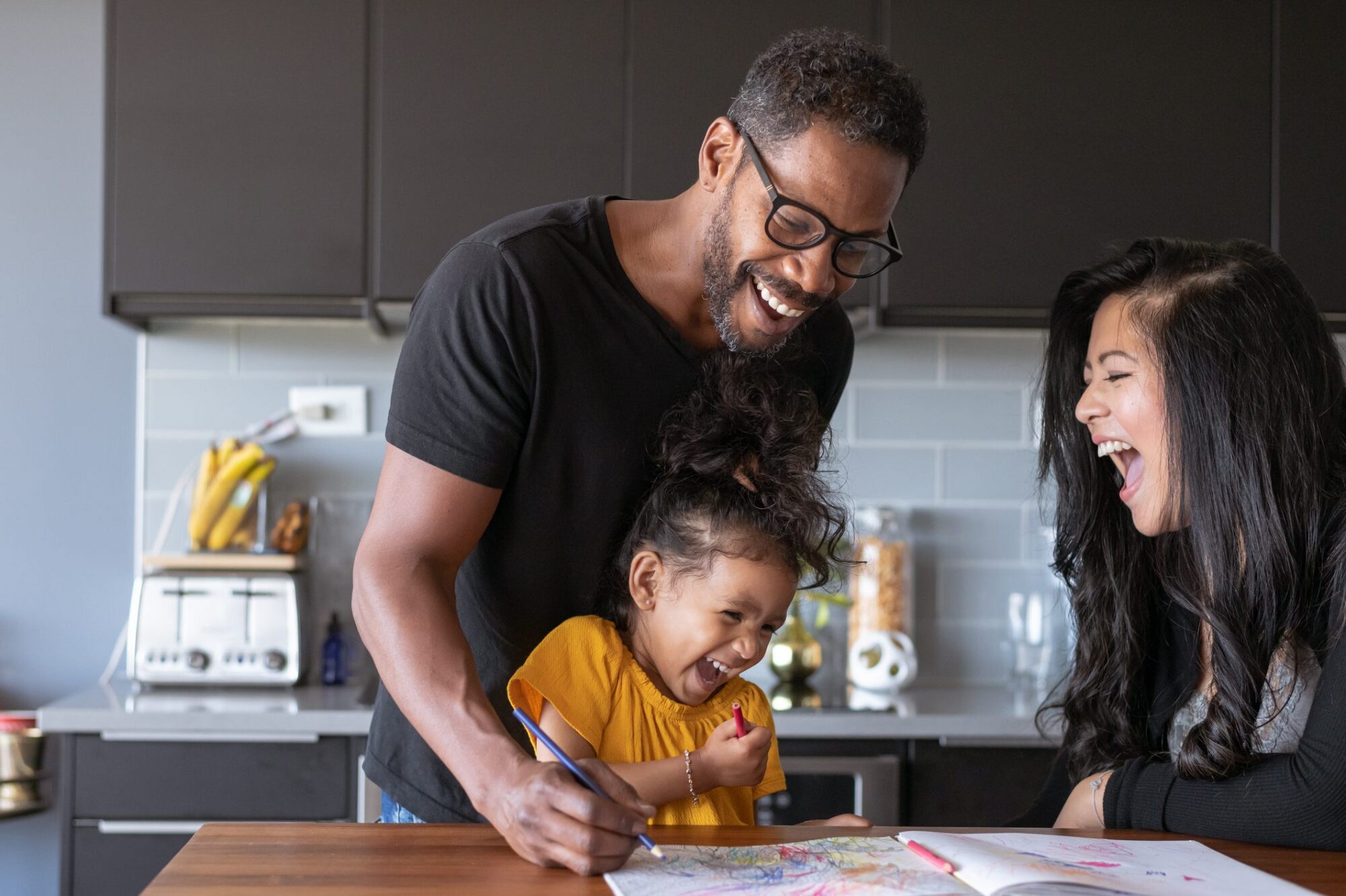 A man and woman smile as a laughing young girl draws with crayons at a kitchen table.