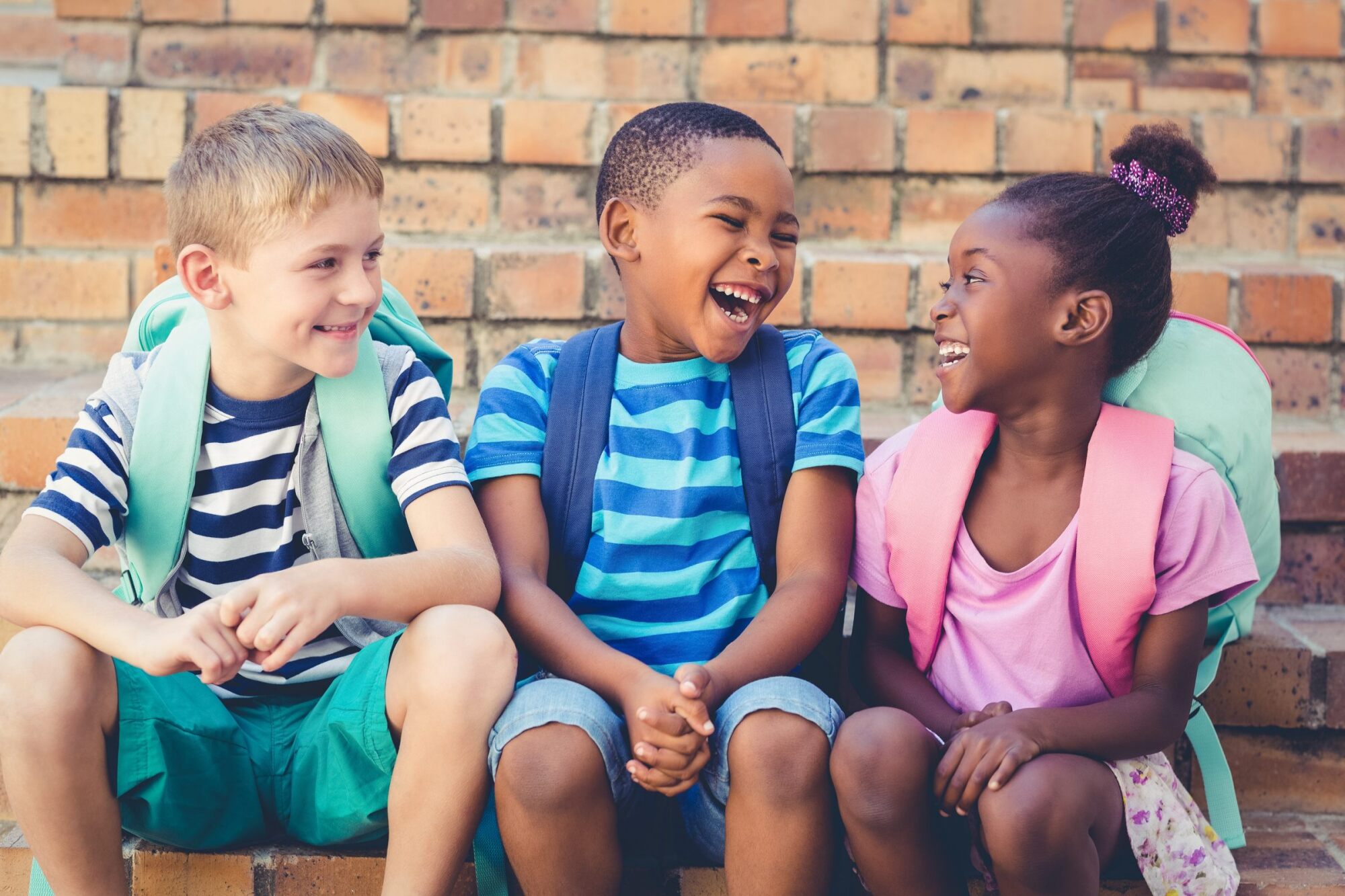 Three children with backpacks sit on steps, laughing and smiling together in front of a brick wall.