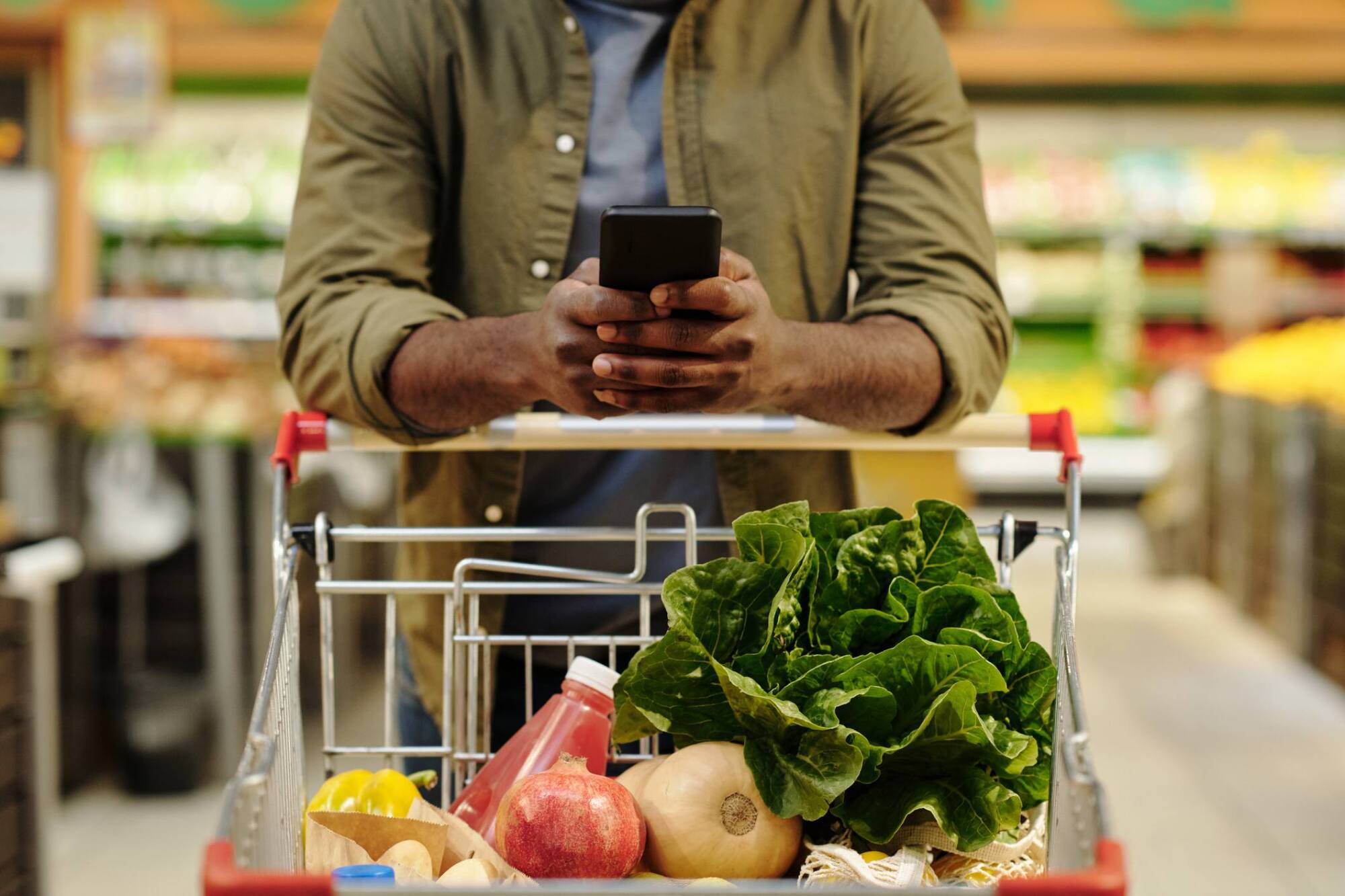 Person using a smartphone while pushing a grocery cart filled with fresh produce in a supermarket.