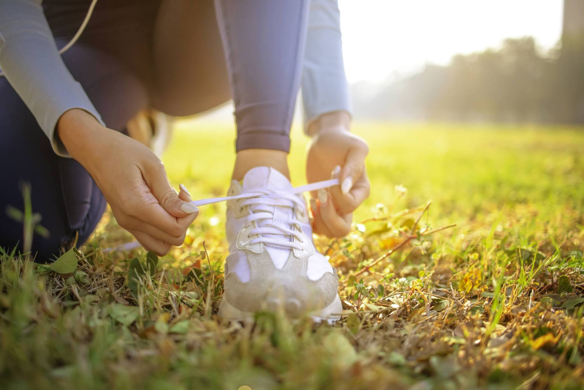 Person kneeling on grass, tying the laces of a white sneaker in bright sunlight.
