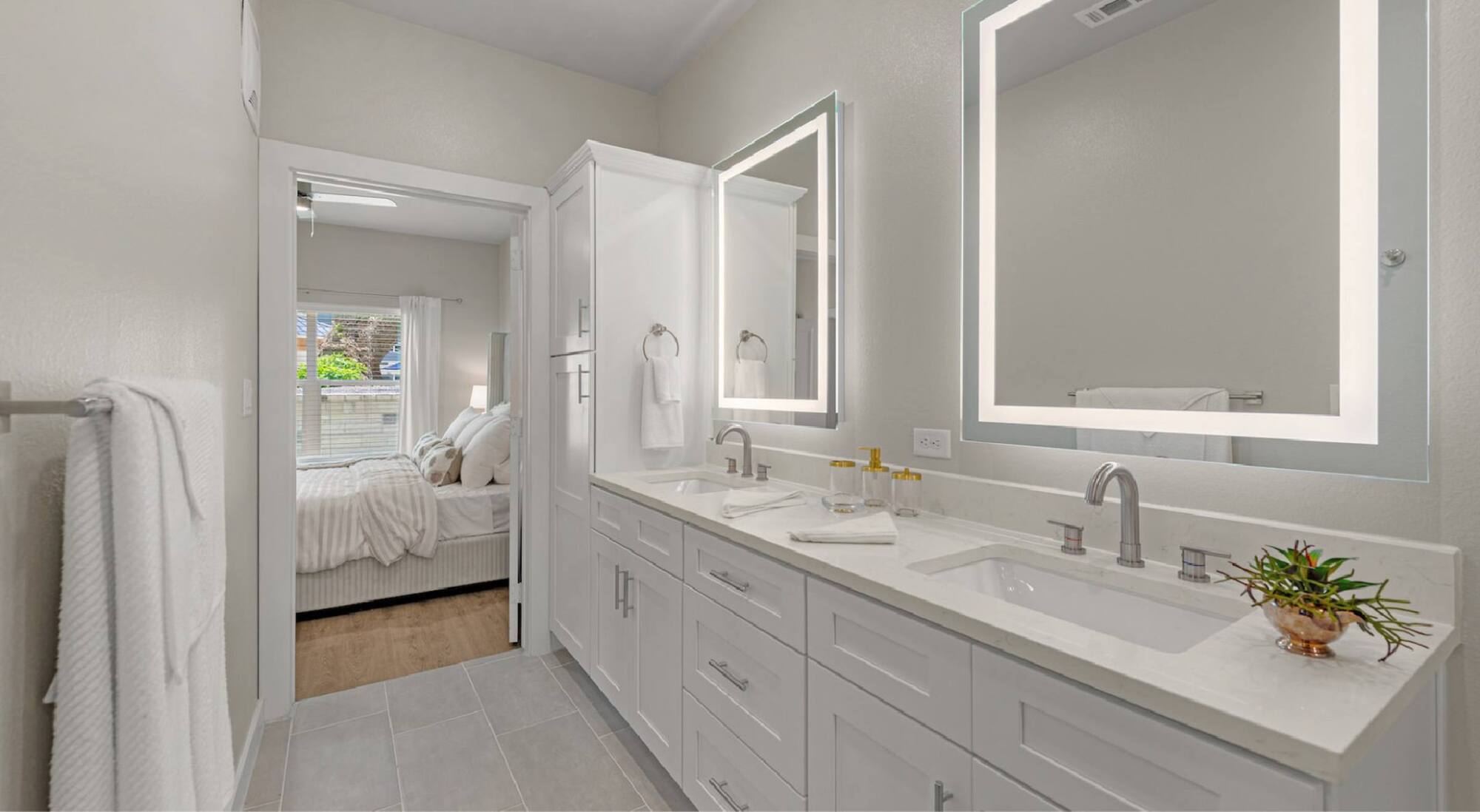 Modern bathroom with double sinks, white cabinets, lighted mirrors, and a view into a bright bedroom.