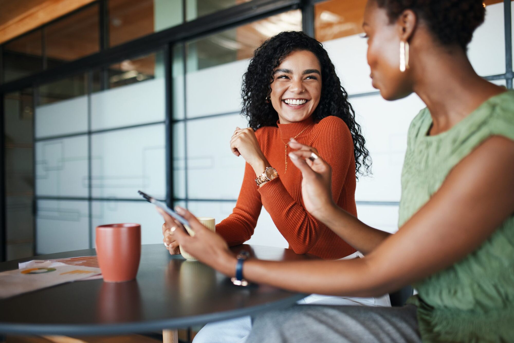 Two women sit at a table, smiling and talking, with coffee cups and a smartphone in front of them.