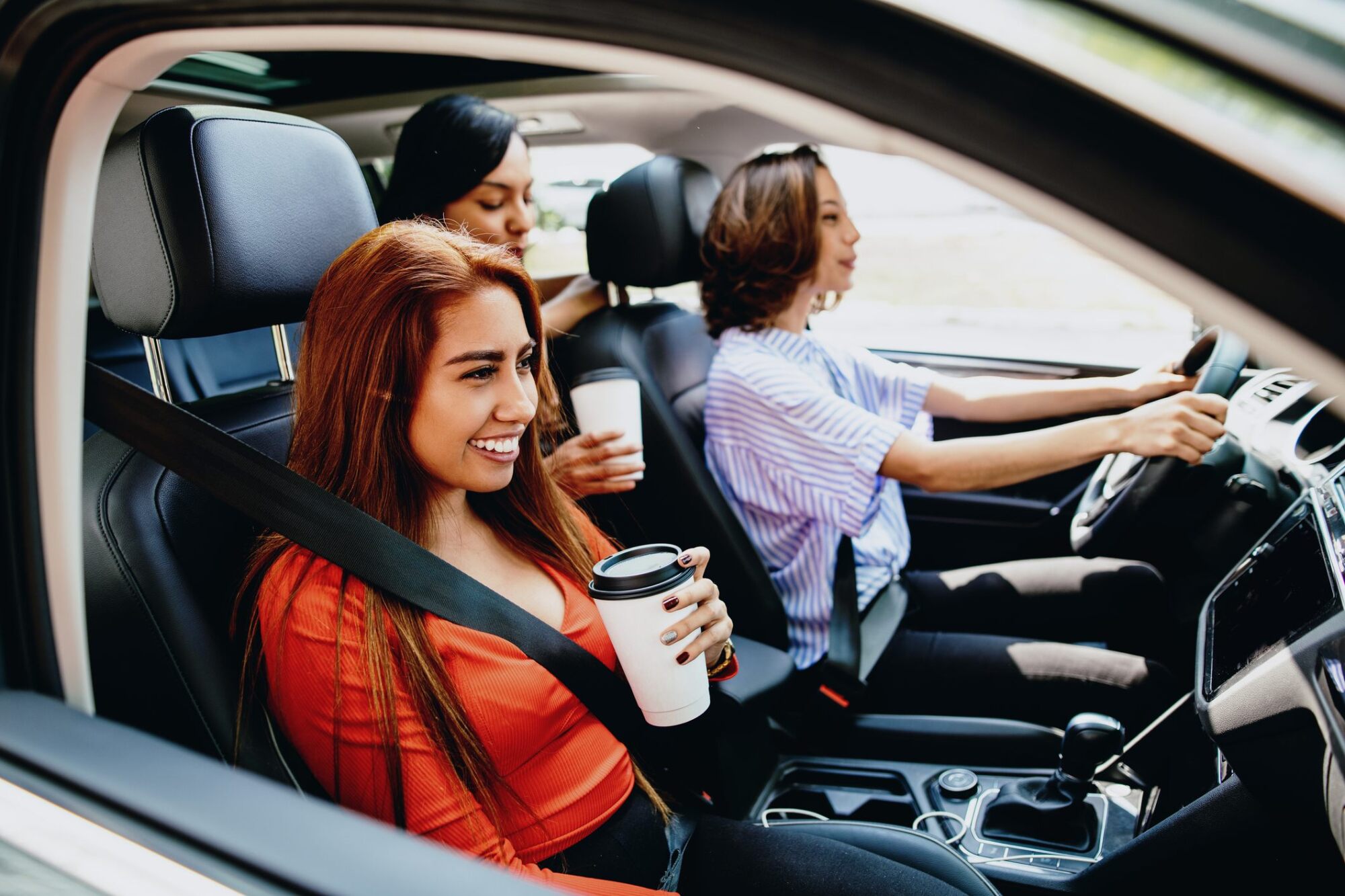 Three young women in a car, smiling and holding coffee cups, with one woman driving.