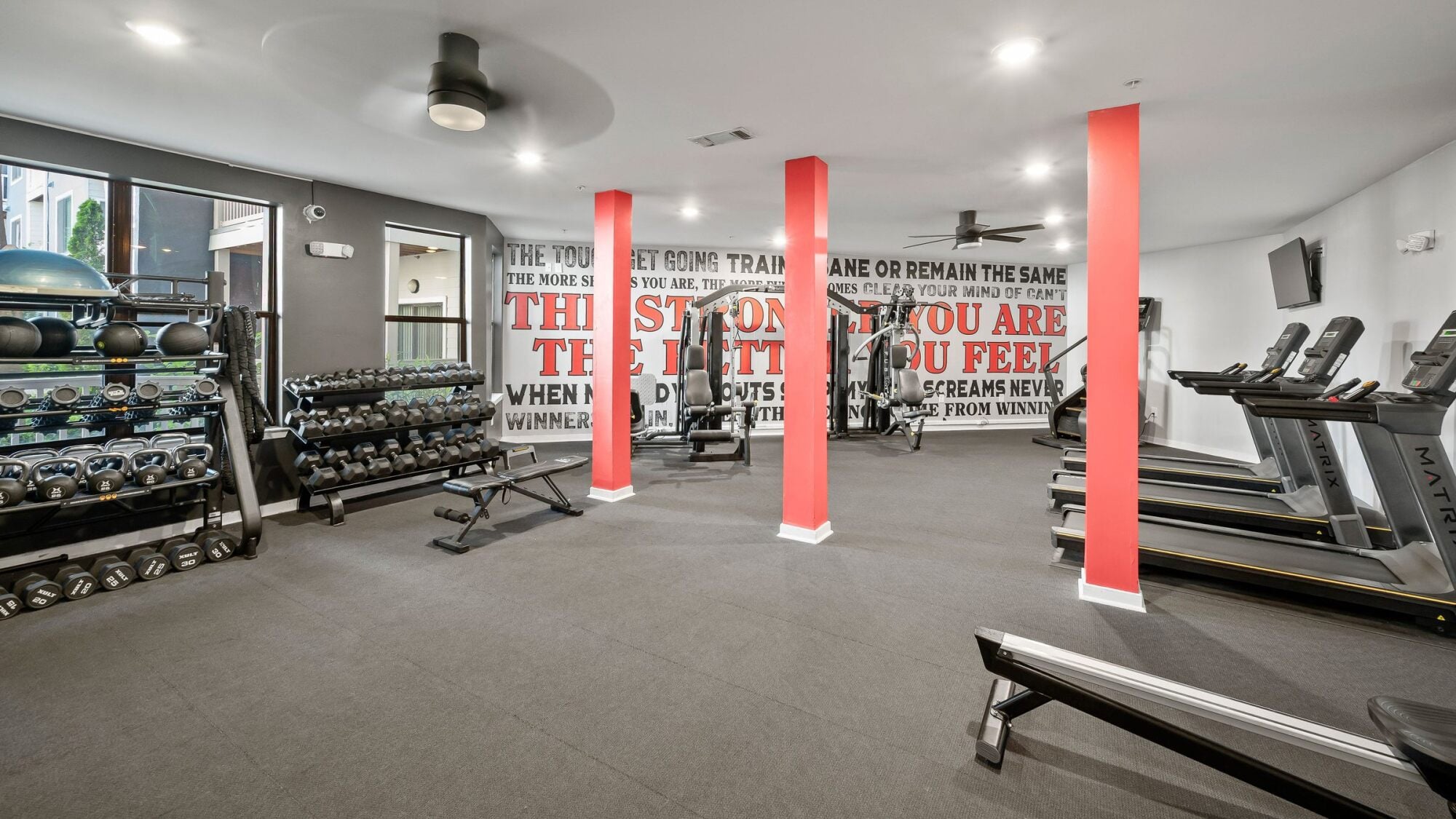 Modern gym room with dumbbells, benches, exercise machines, red columns, and motivational wall text at Apex West Midtown.