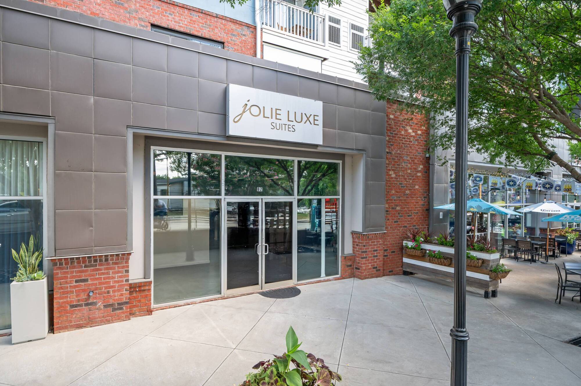 Street view of Jolie Luxe Suites entrance with glass doors, sign above, and plants in front.
