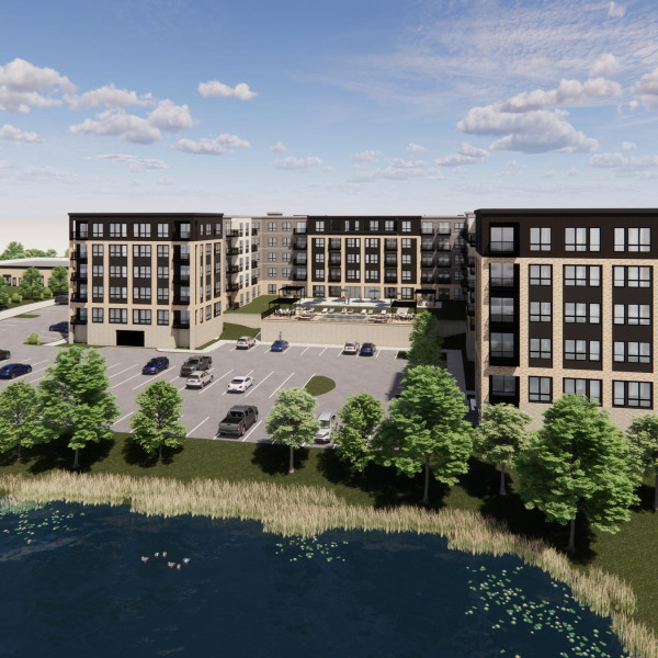 Modern apartment complex with parking lot, trees, and a pond in the foreground under a partly cloudy sky.