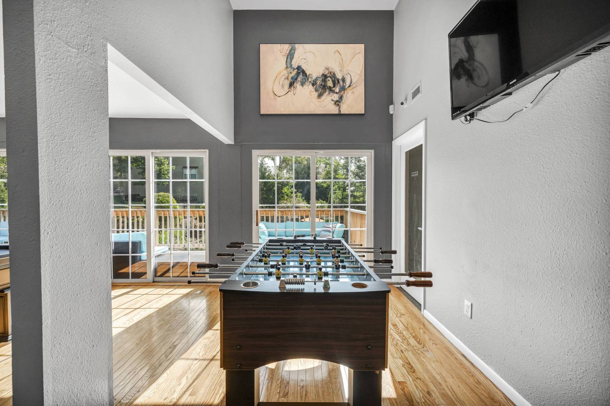 A foosball table in a bright room with large windows, wood floors, and a TV mounted on a gray wall.