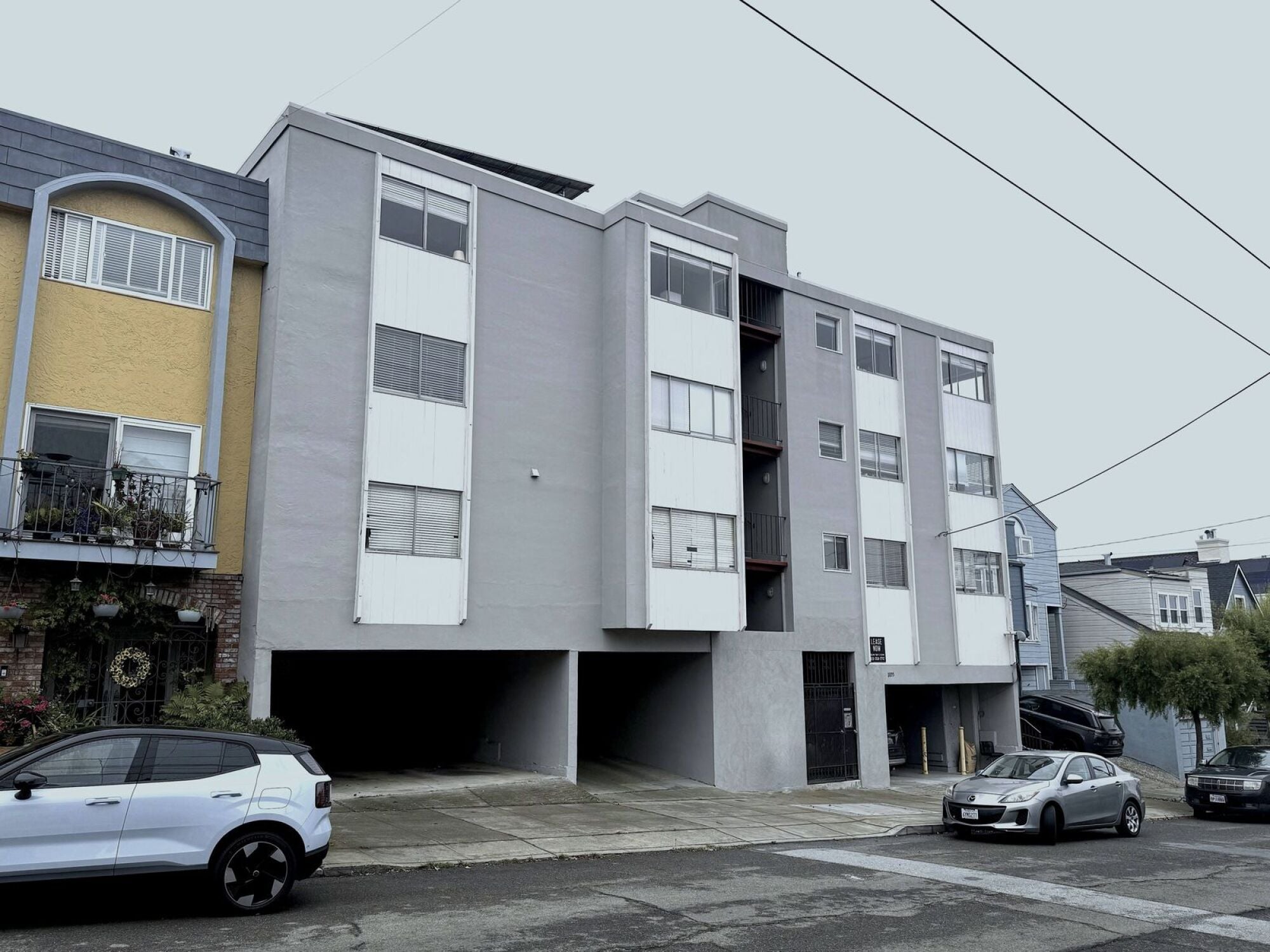 A white car is parked in front of a grey and yellow apartment building.