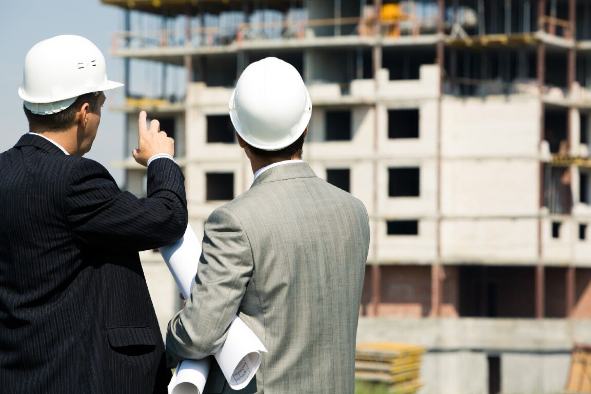 Two men in suits and white hard hats discuss blueprints at a construction site with a building in progress.