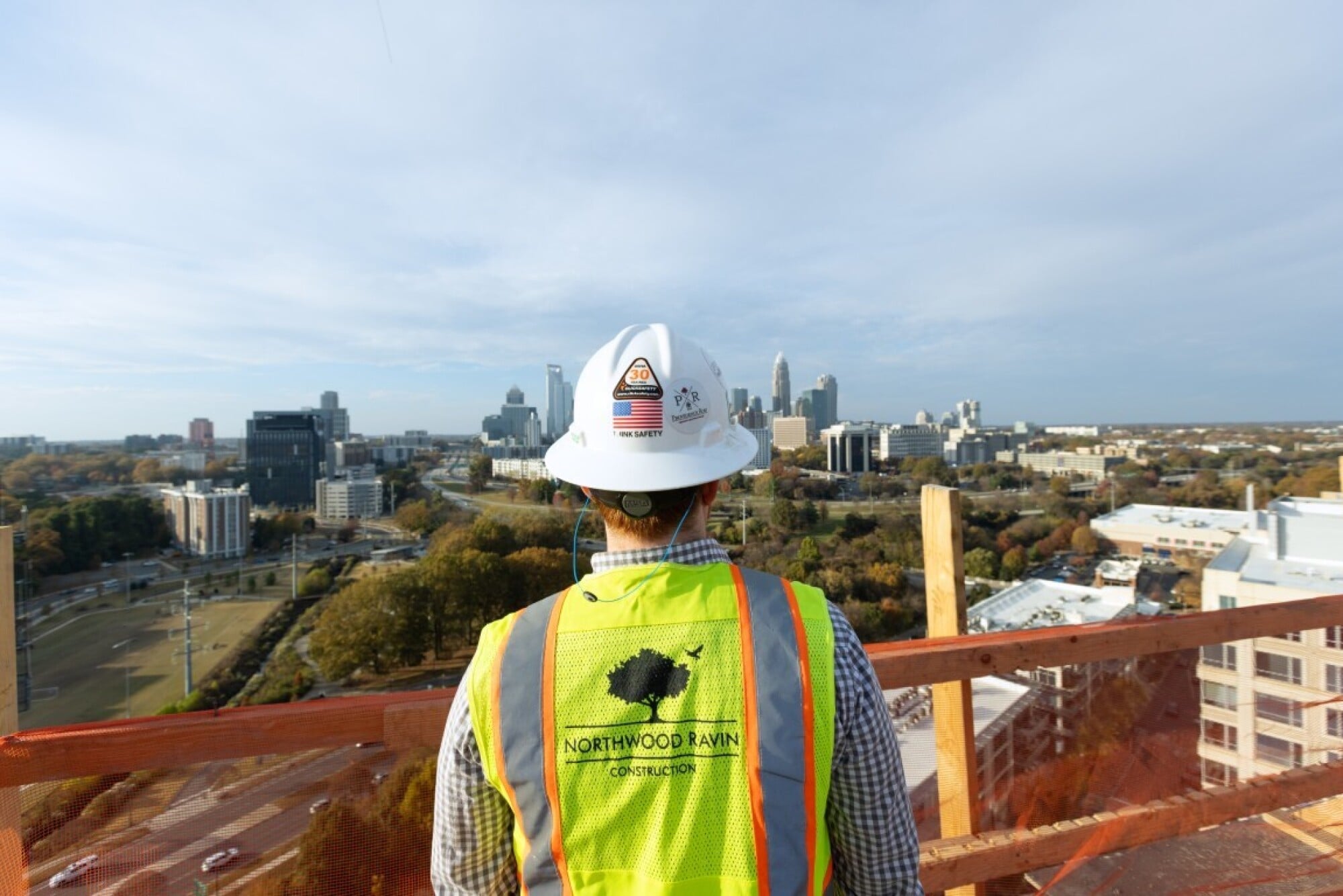 Construction worker in safety gear overlooks a city skyline from a high rooftop on a clear day.