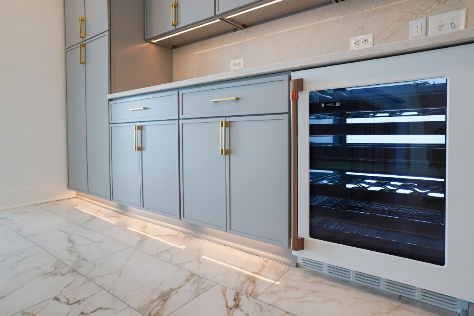 Modern kitchen with blue cabinets, gold handles, marble floor, and a built-in glass-front wine fridge under the countertop.