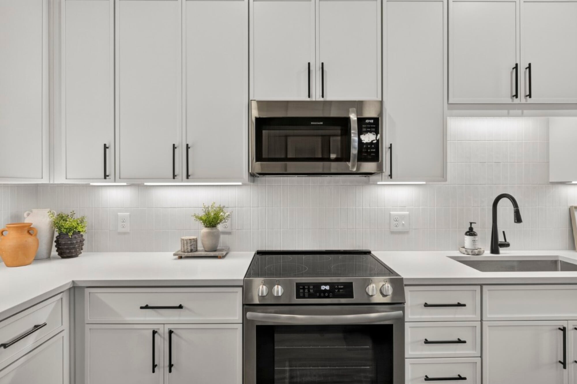 Modern white kitchen with stainless steel oven, microwave, black faucet, and decorative plants on white countertops.