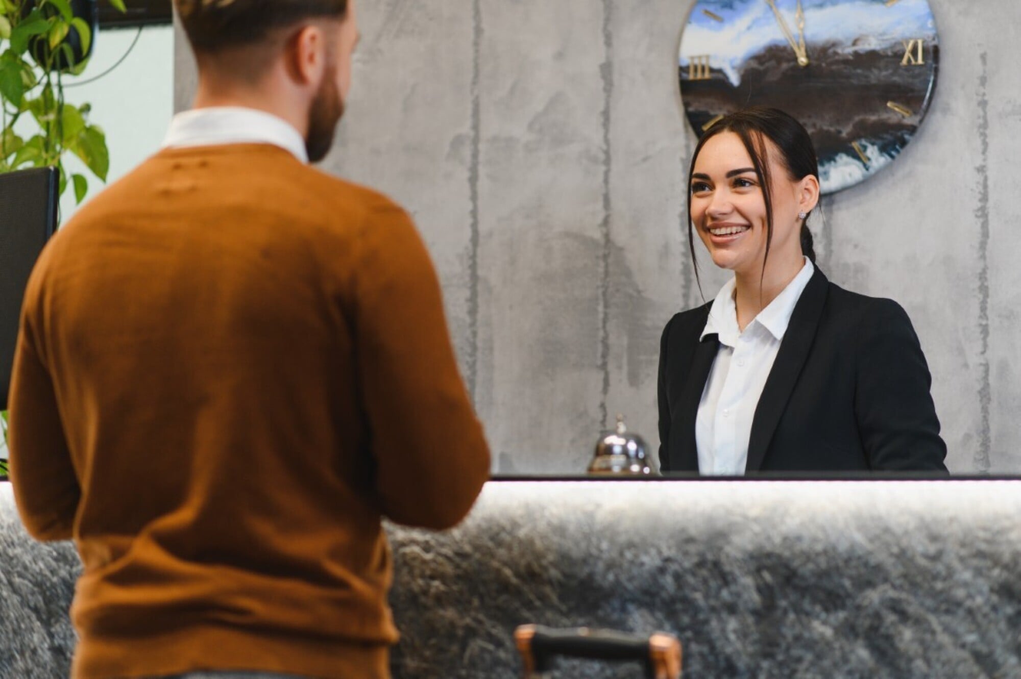 A smiling receptionist in a black blazer greets a man at a hotel front desk with a clock on the wall behind her.