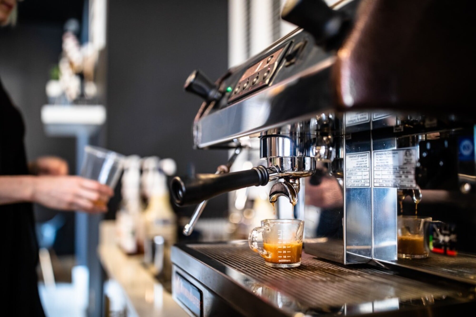 Espresso machine brewing coffee into a glass cup, with a person holding a cup in the blurred background.