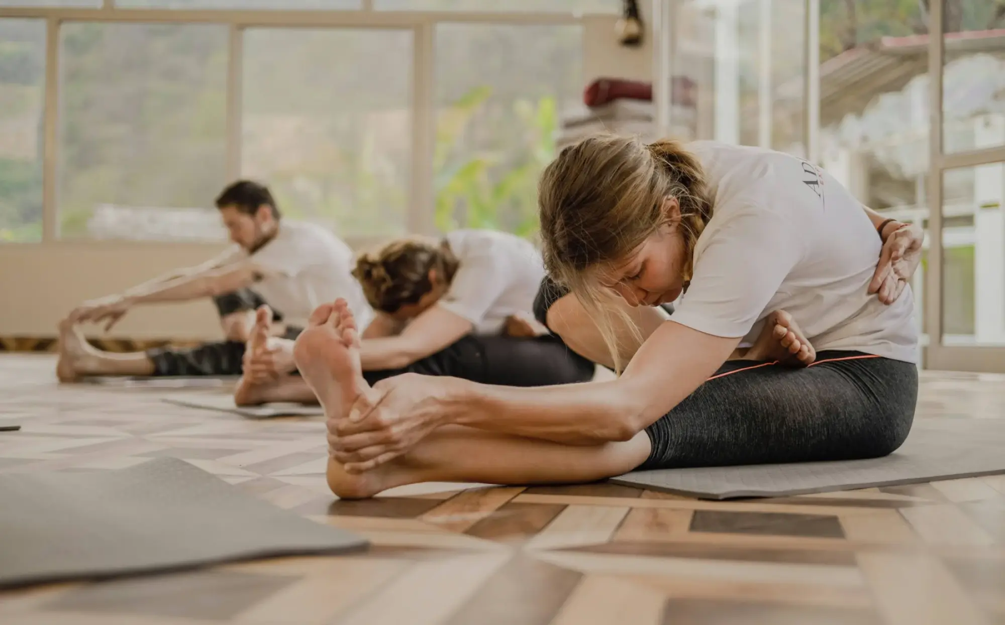People in a bright room doing seated yoga stretches on mats, reaching for their feet with concentration.