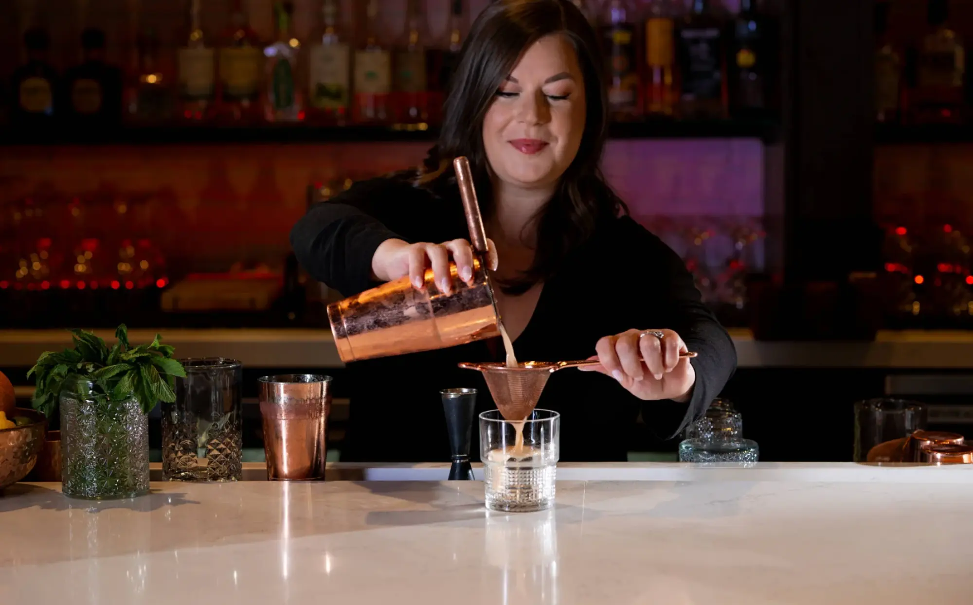 A woman behind a bar pours a drink through a strainer into a glass, smiling as she works. Bottles are visible on shelves in the background.