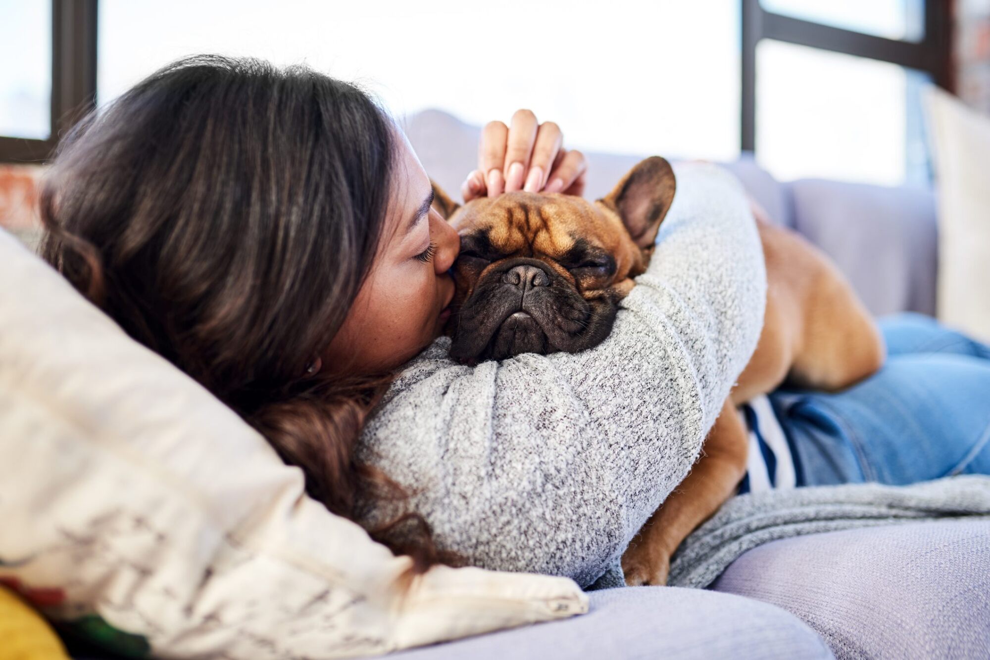 Woman lying on a couch hugging and kissing a brown French bulldog, both looking relaxed and cozy.