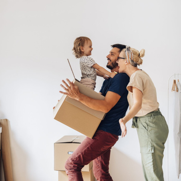 A smiling family carries moving boxes in a new home, with a child laughing in a parent's arms.
