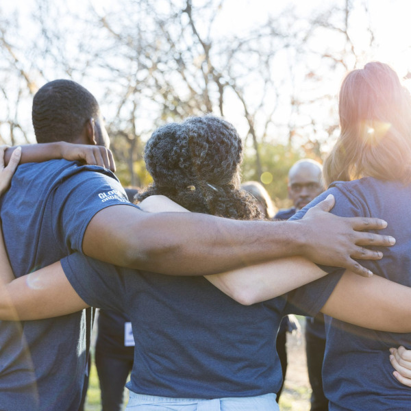 A group of people in blue shirts stand outdoors, arms around each other in a circle, facing away.