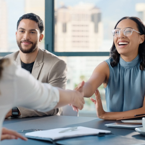 Smiling woman in glasses shakes hands with another person across a table during a meeting.