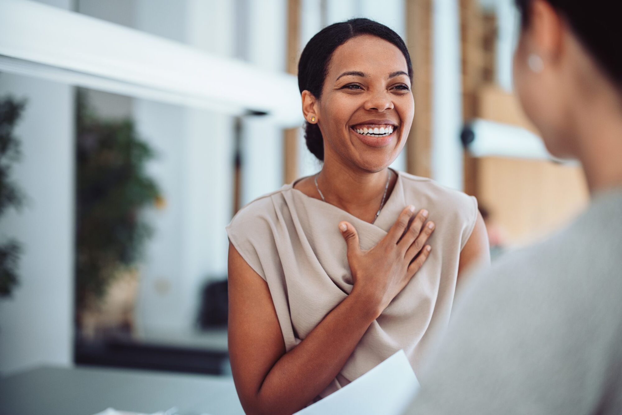 Smiling woman with hand on chest talks to another person indoors.