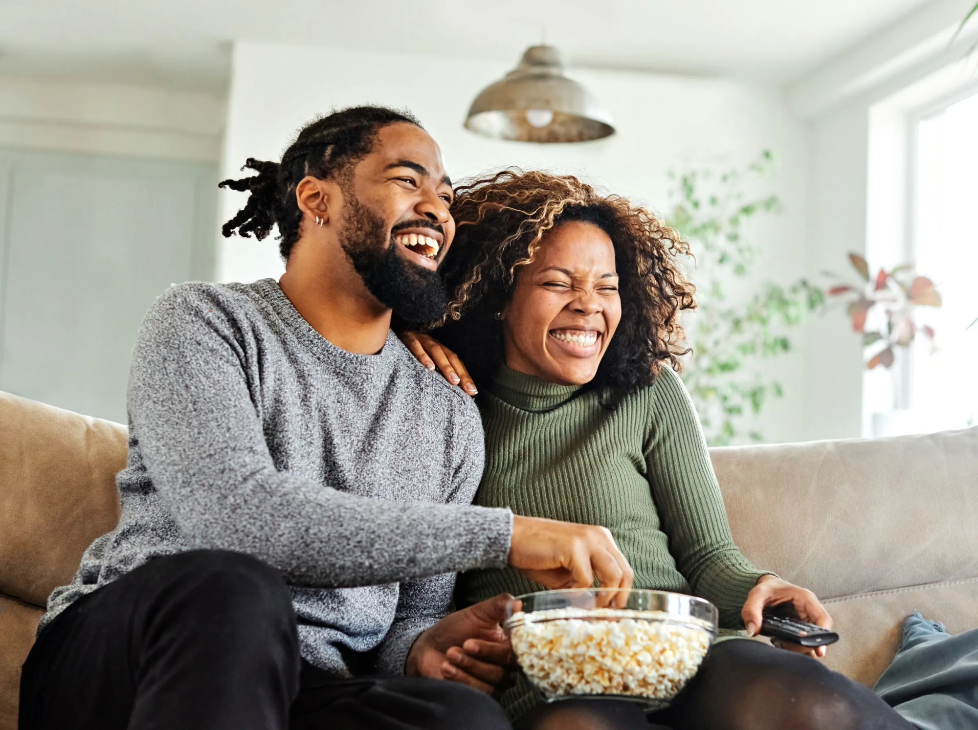 Smiling couple sitting on a couch, eating popcorn, and watching TV together in a bright living room.