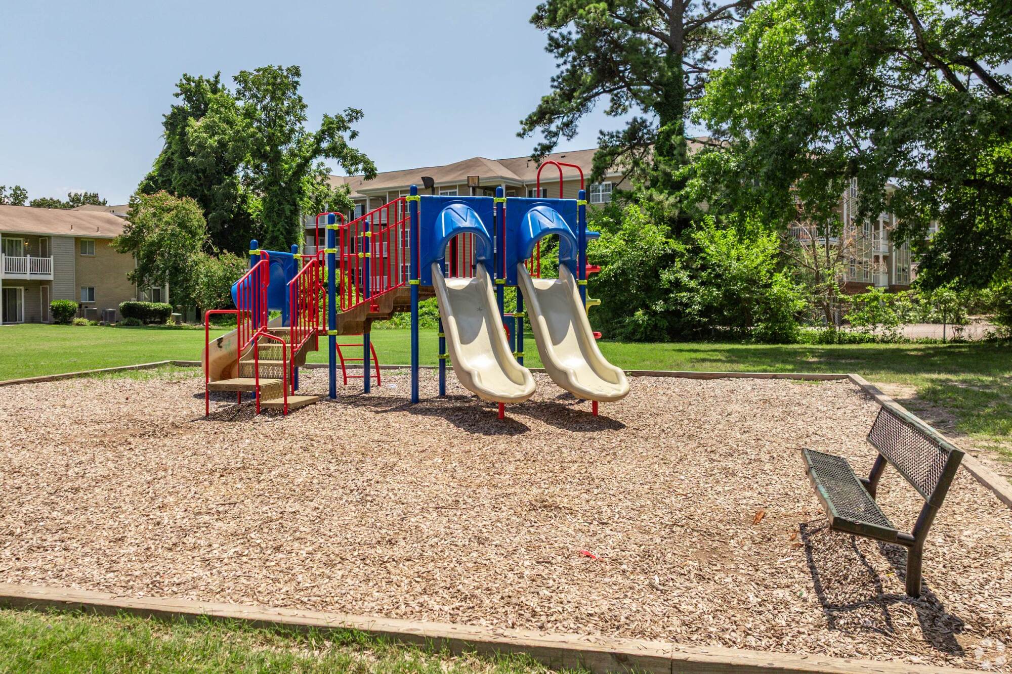 Playground with two slides, stairs, and wood chips; surrounded by grass, trees, and apartment buildings, with a bench in front.