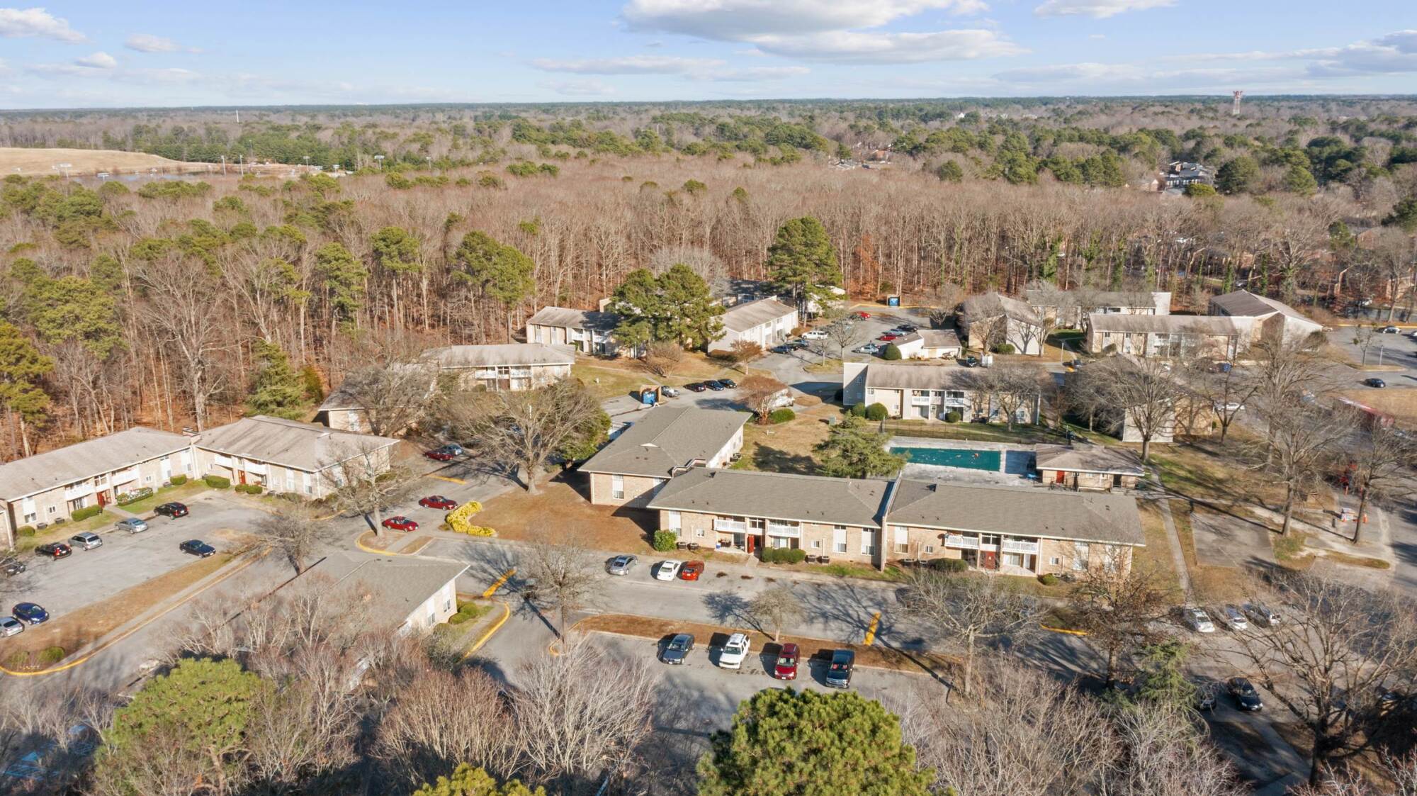 Aerial view of apartment buildings surrounded by trees, parking lots, and cars on a clear day.