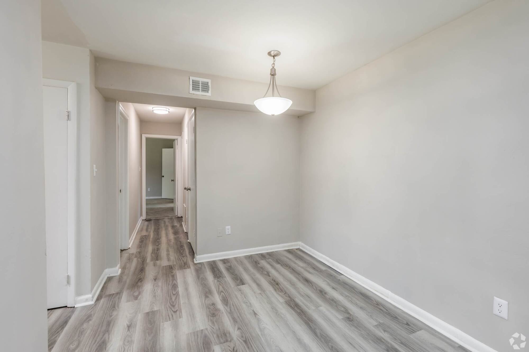 Empty room with light gray walls, wood-look flooring, ceiling light fixture, and hallway leading to doorways.