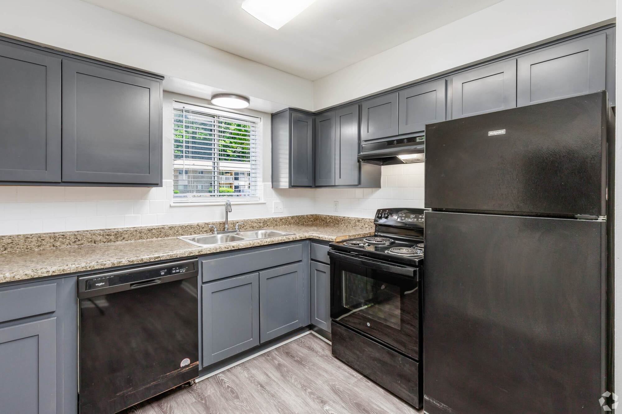 Modern kitchen with gray cabinets, black appliances, double sink, granite countertops, and a window with blinds above the sink.