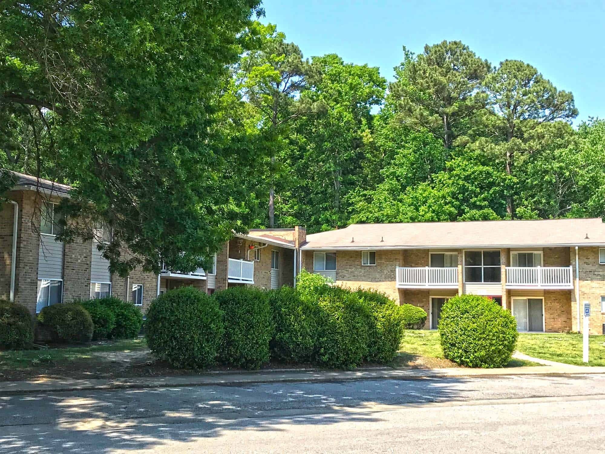 Two-story apartment buildings with balconies, surrounded by green trees and shrubs on a sunny day.