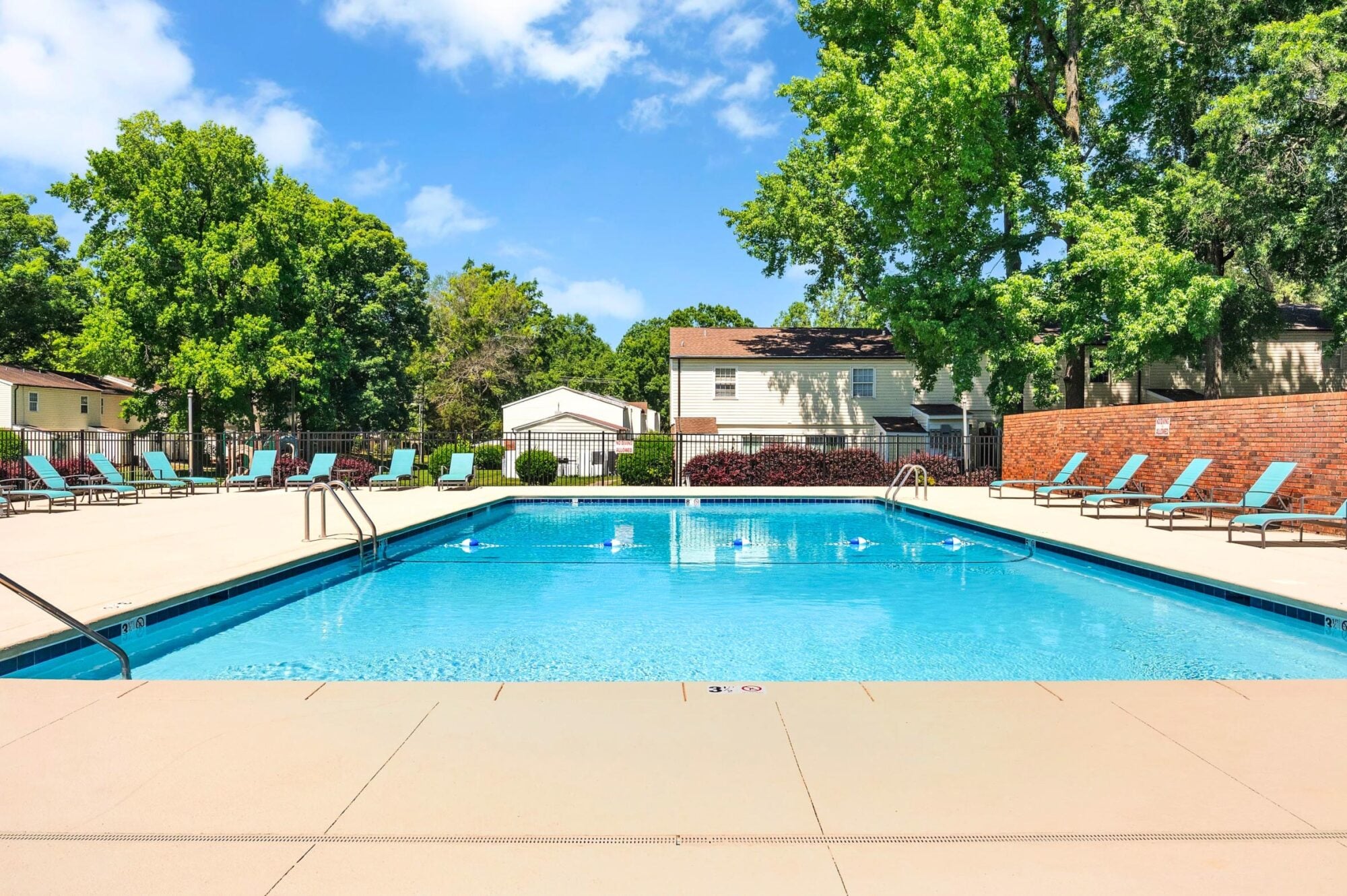 Outdoor swimming pool with lounge chairs, surrounded by trees and residential buildings under a blue sky.