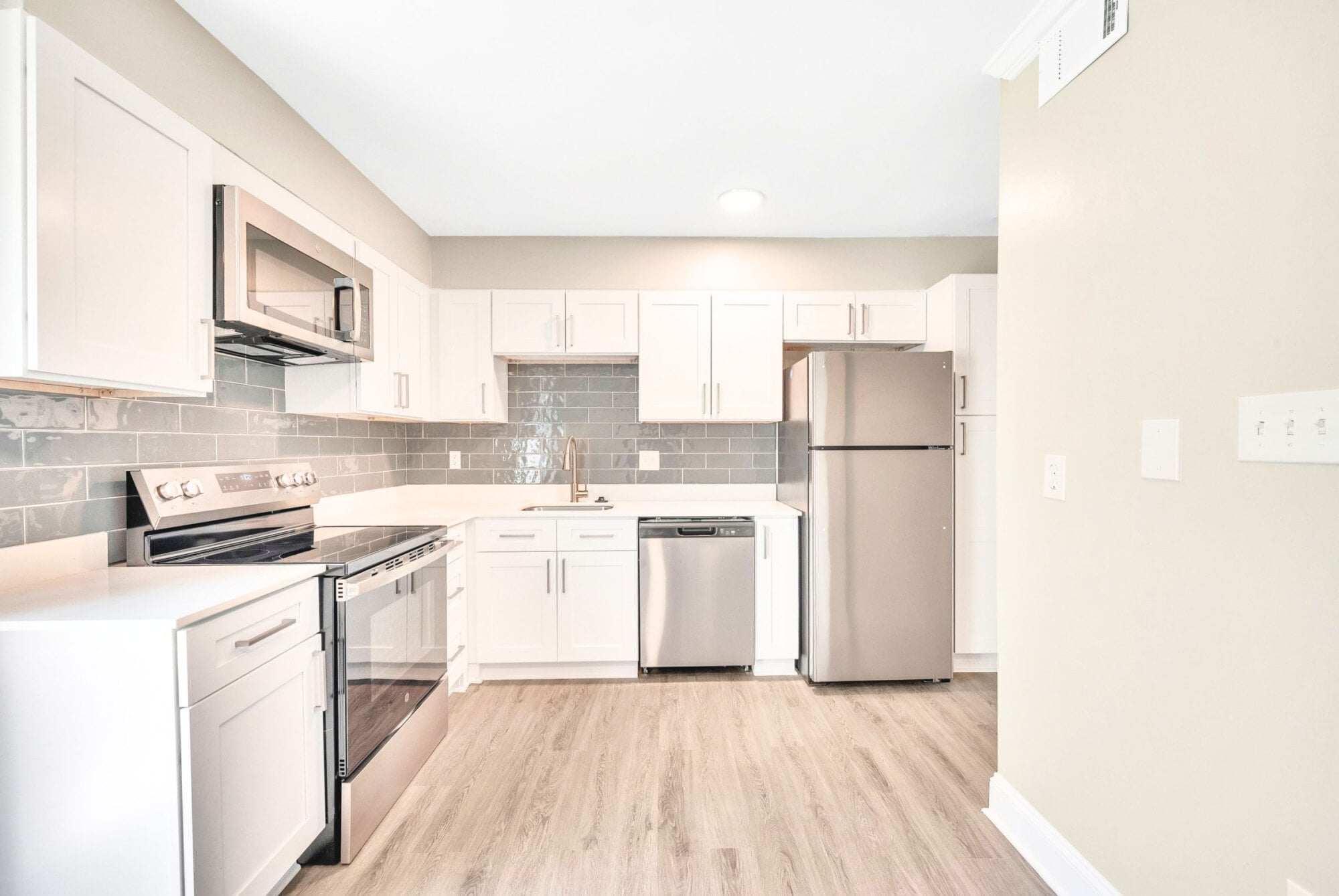 Bright modern kitchen with white cabinets, stainless steel appliances, gray tile backsplash, and light wood flooring.