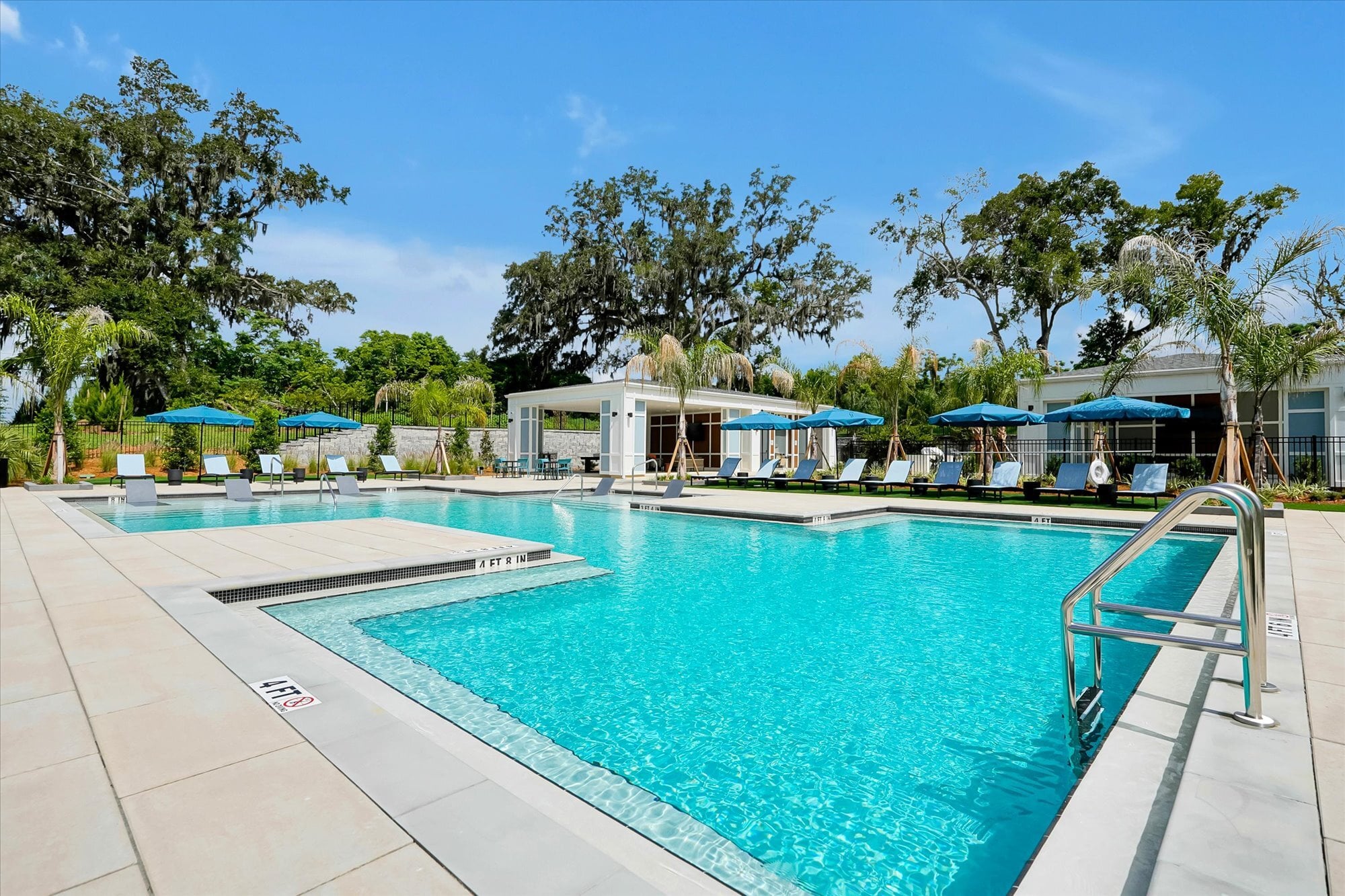 Outdoor swimming pool with clear blue water, lounge chairs, and blue umbrellas, surrounded by trees on a sunny day.