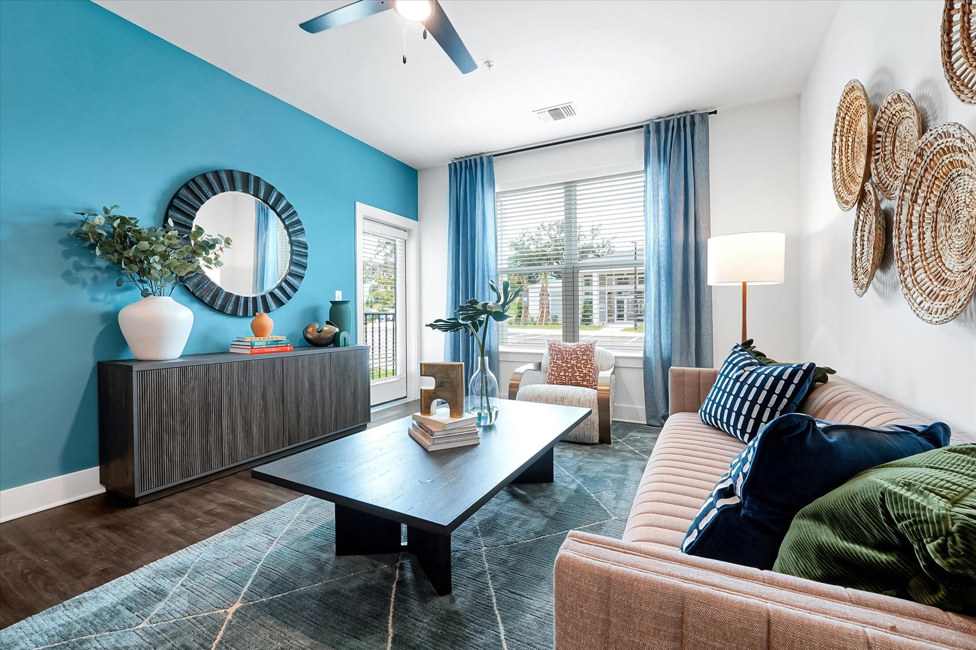 Modern living room with a blue accent wall, tan sofa, coffee table, and large window with blue curtains letting in natural light.