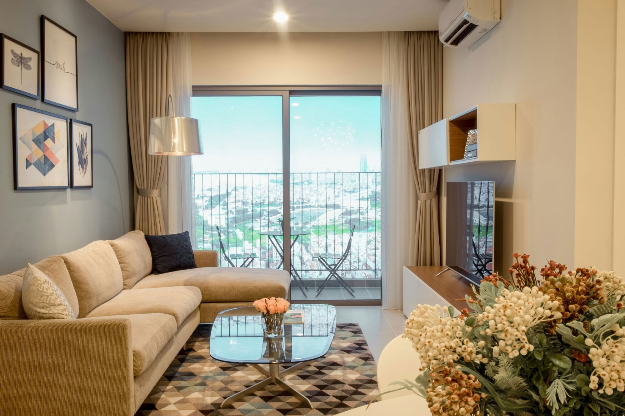 Modern living room with beige sofa, glass coffee table, TV, balcony view, and decorative wall art and flowers.
