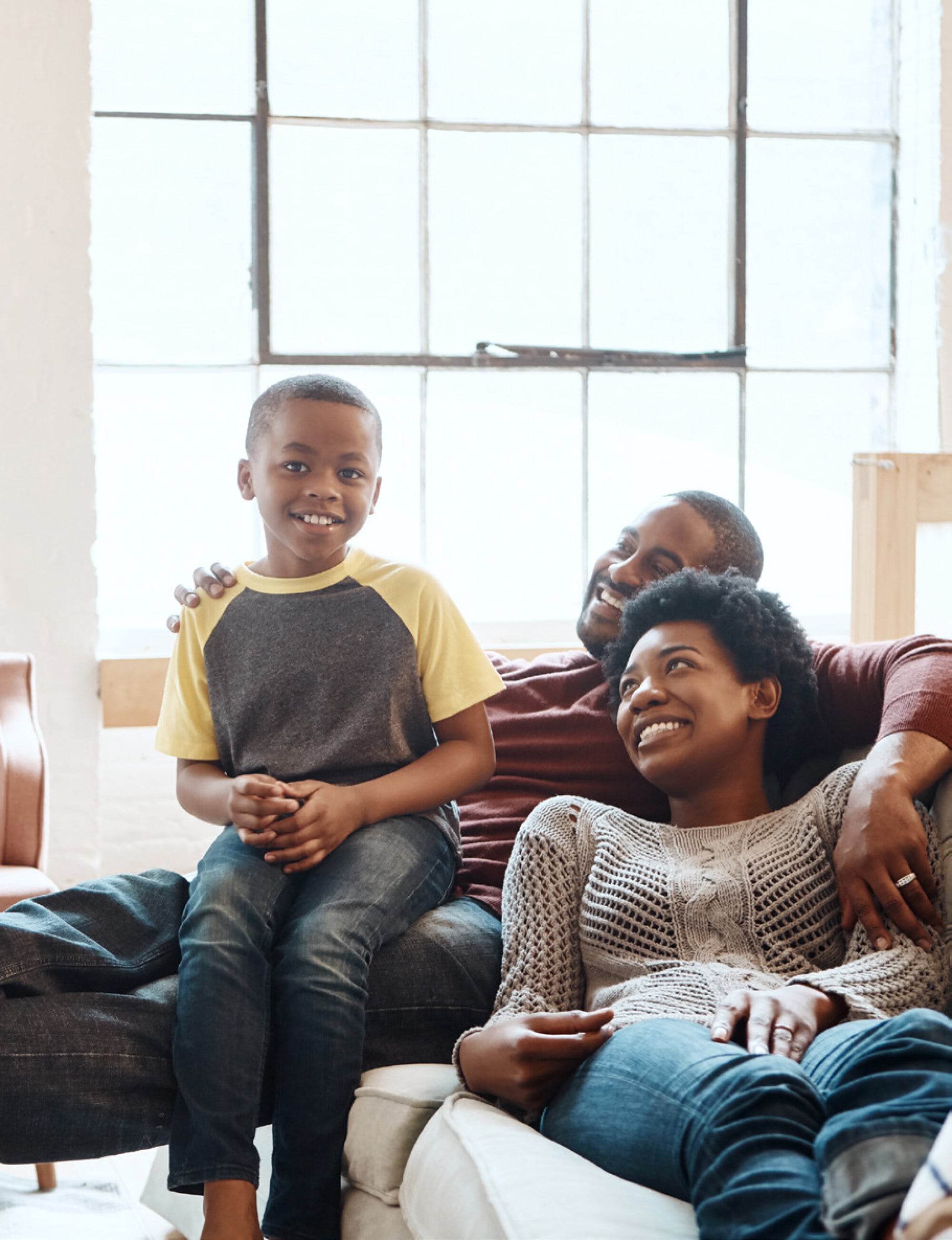 A smiling family of three relaxes together on a couch in a bright living room with large windows behind them.