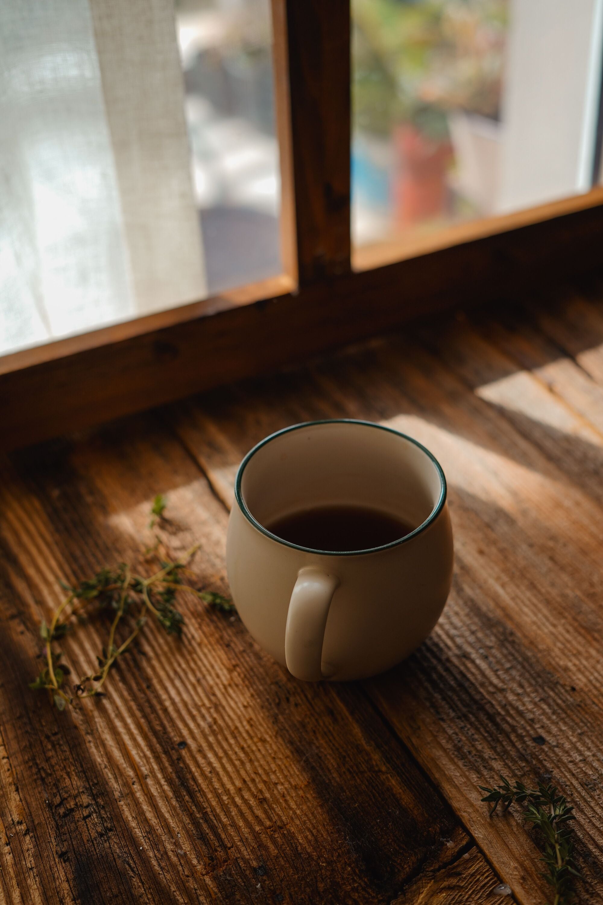 A white mug of tea sits on a rustic wooden table by a sunlit window with a sheer curtain and some green herbs nearby.