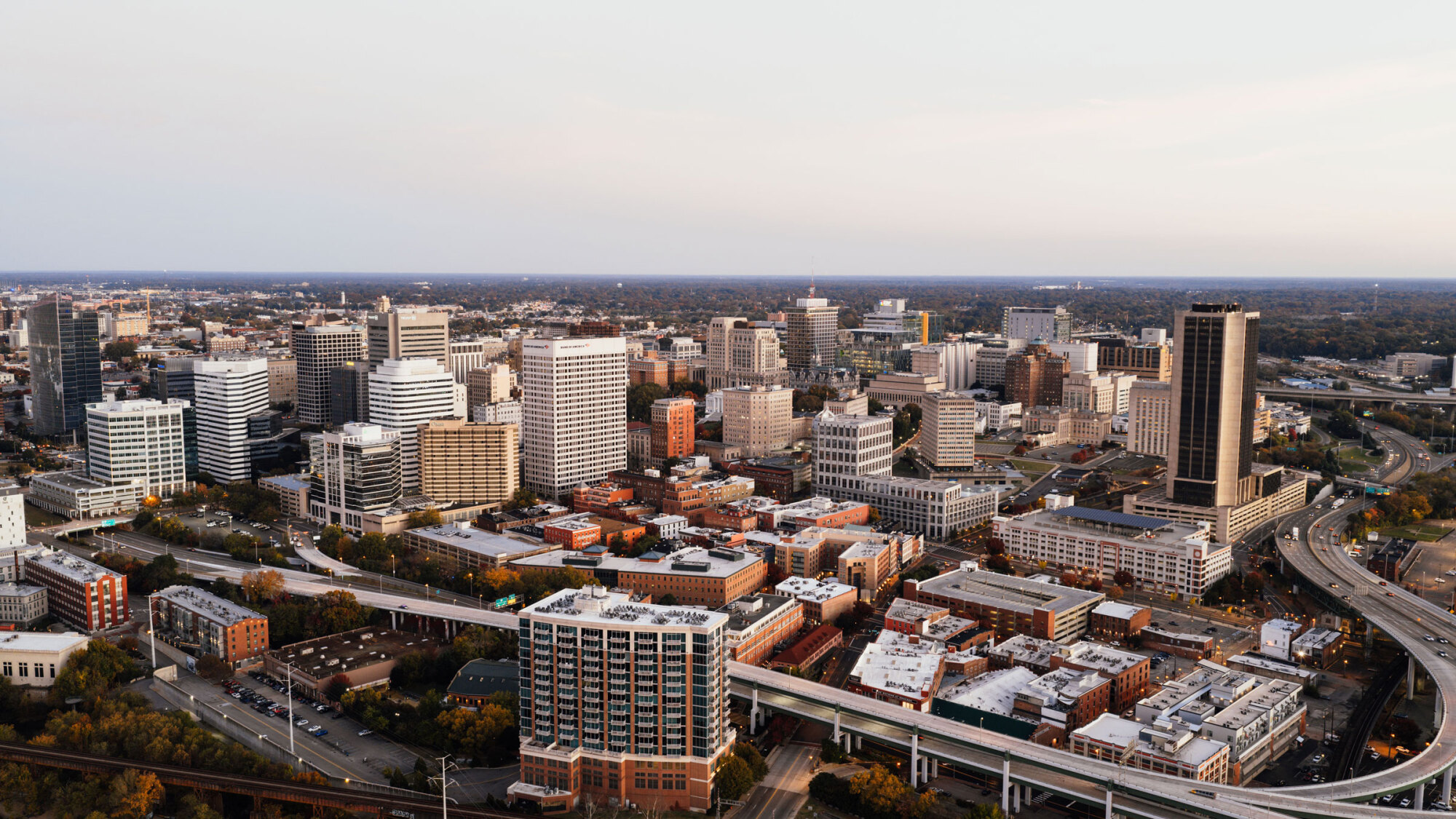 Aerial view of a city skyline with tall buildings, highways, and scattered trees under a clear sky.