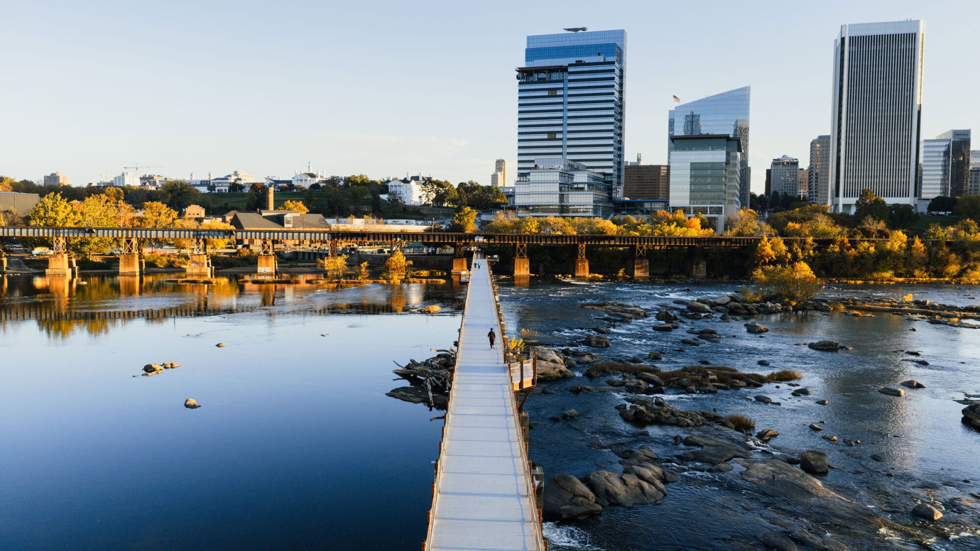 Pedestrian bridge over a river with rocks, leading to a city skyline and modern buildings in the background.