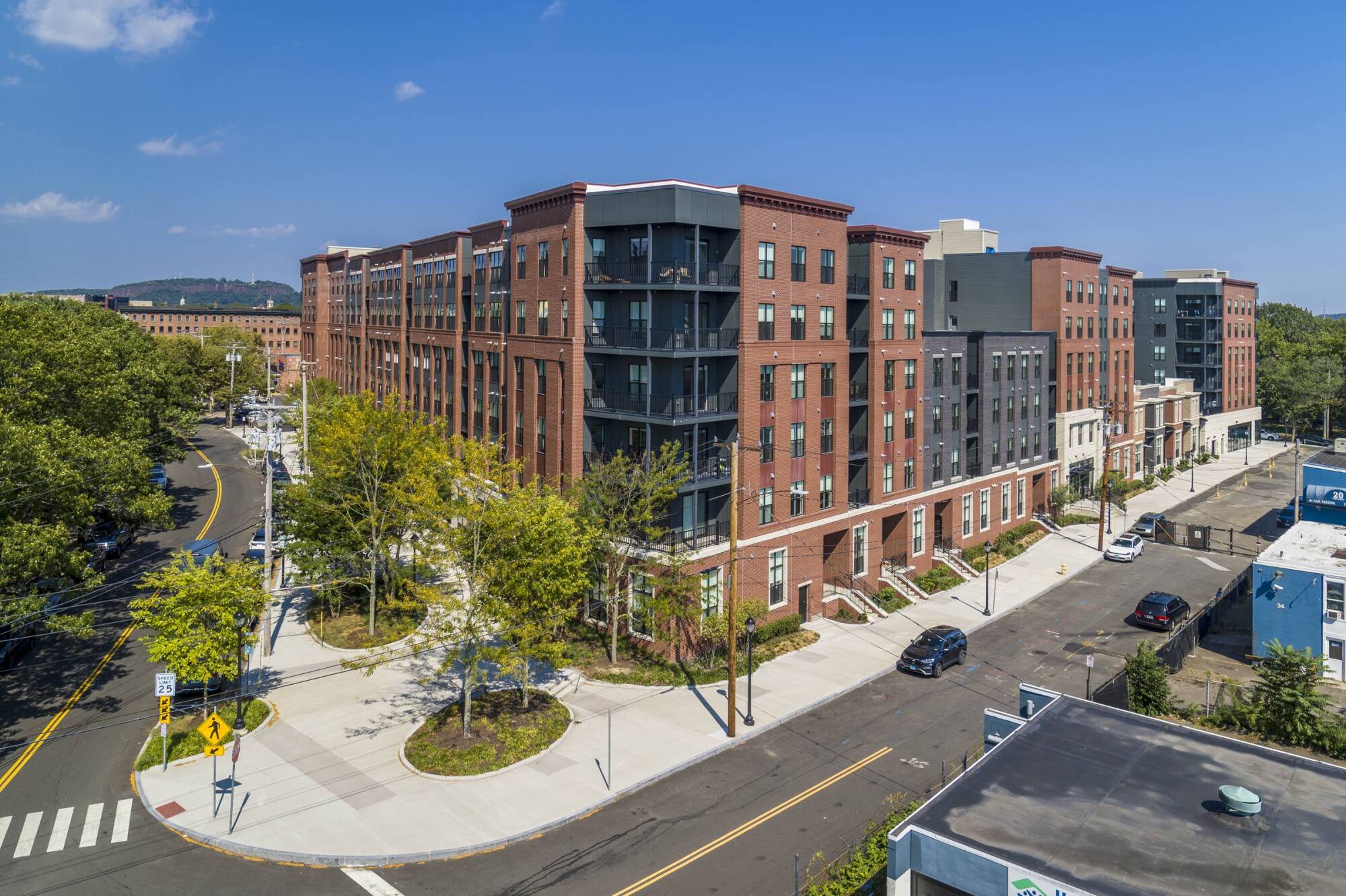 A red-brick apartment building on a sunny day, with trees, parked cars, and a sidewalk at a street corner.