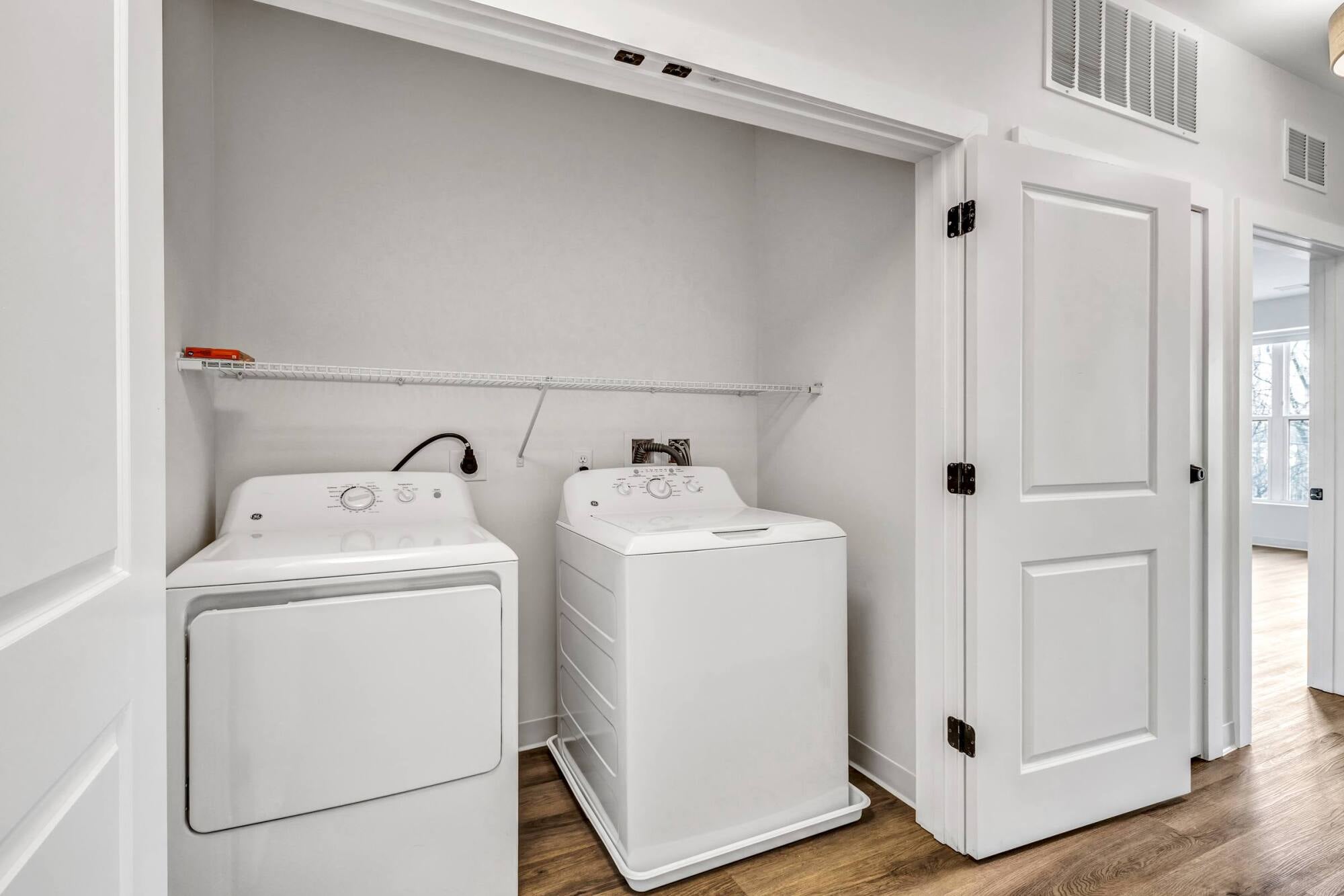 A laundry closet with a white washer and dryer, a wire shelf above, and double doors open to a hallway with wooden flooring.