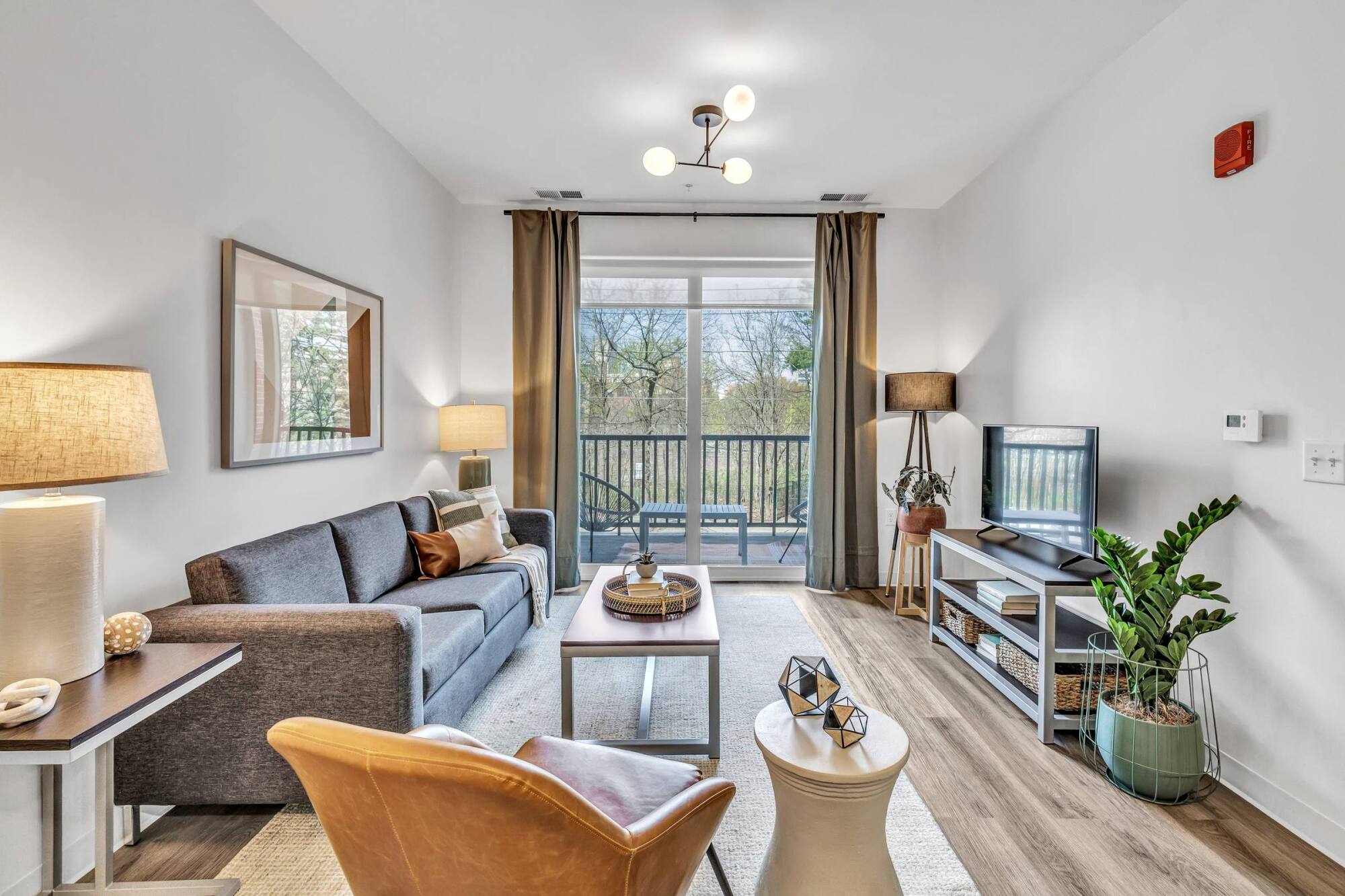 Modern living room with sofa, armchair, TV, lamps, coffee table, and large window with balcony view and trees outside.