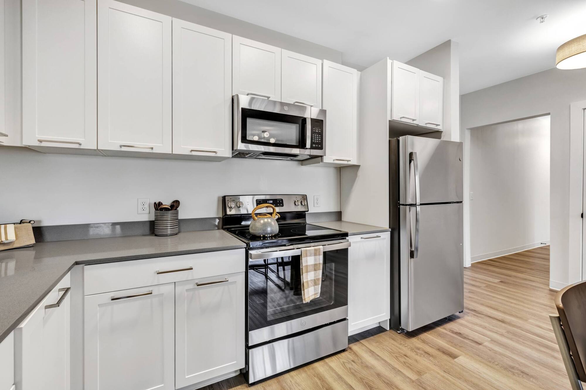 Modern kitchen with white cabinets, stainless steel appliances, and a kettle on the stove. Light wood floor and neutral decor.