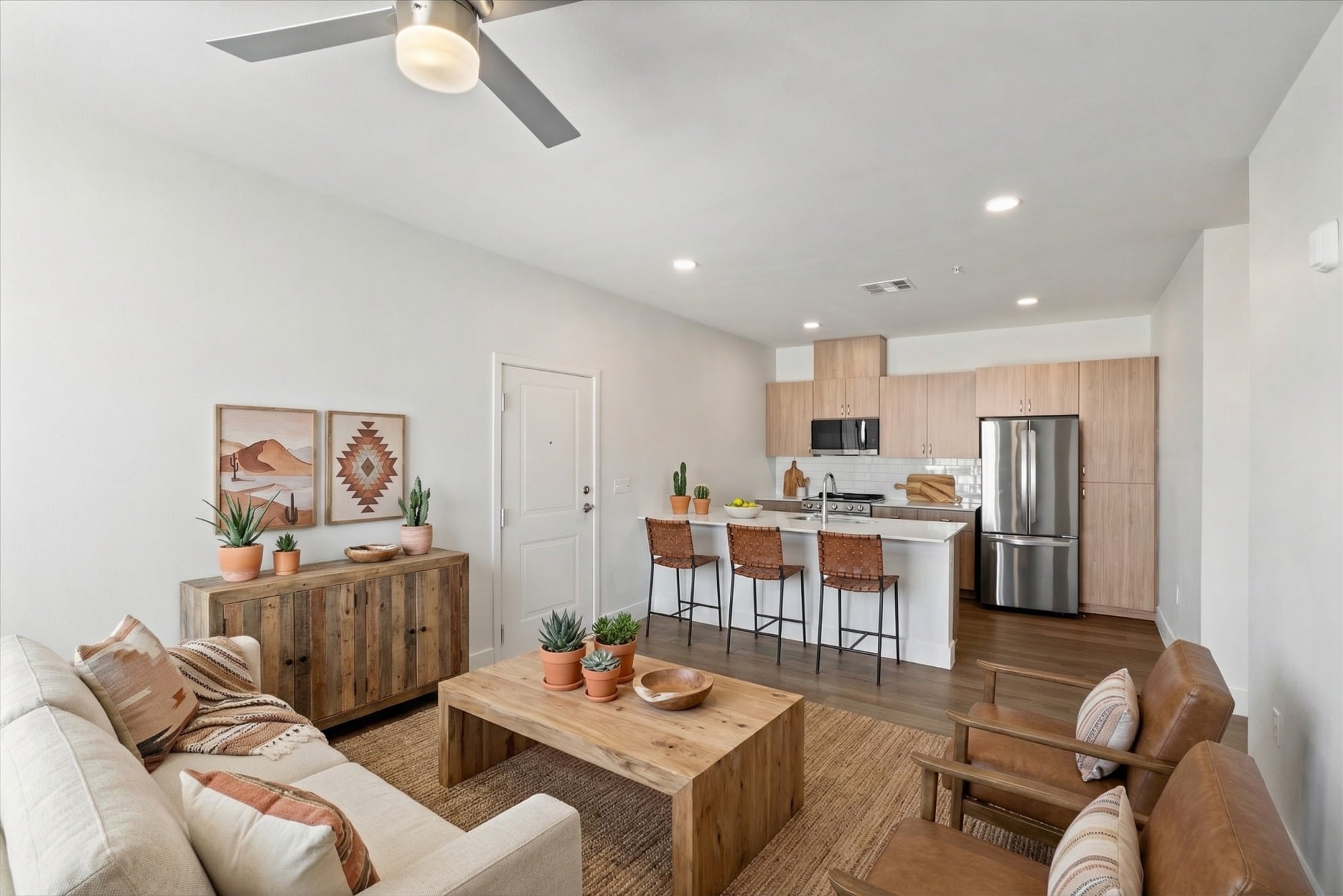 Modern open-concept living room and kitchen with neutral decor, wooden furniture, and potted plants.