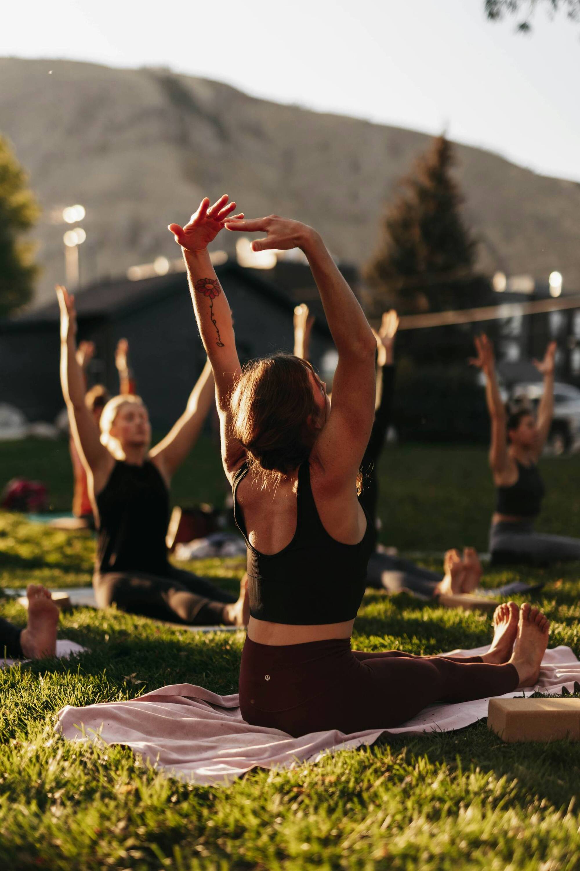 People practicing yoga outdoors on grass, stretching arms upward, with mountains in the background.