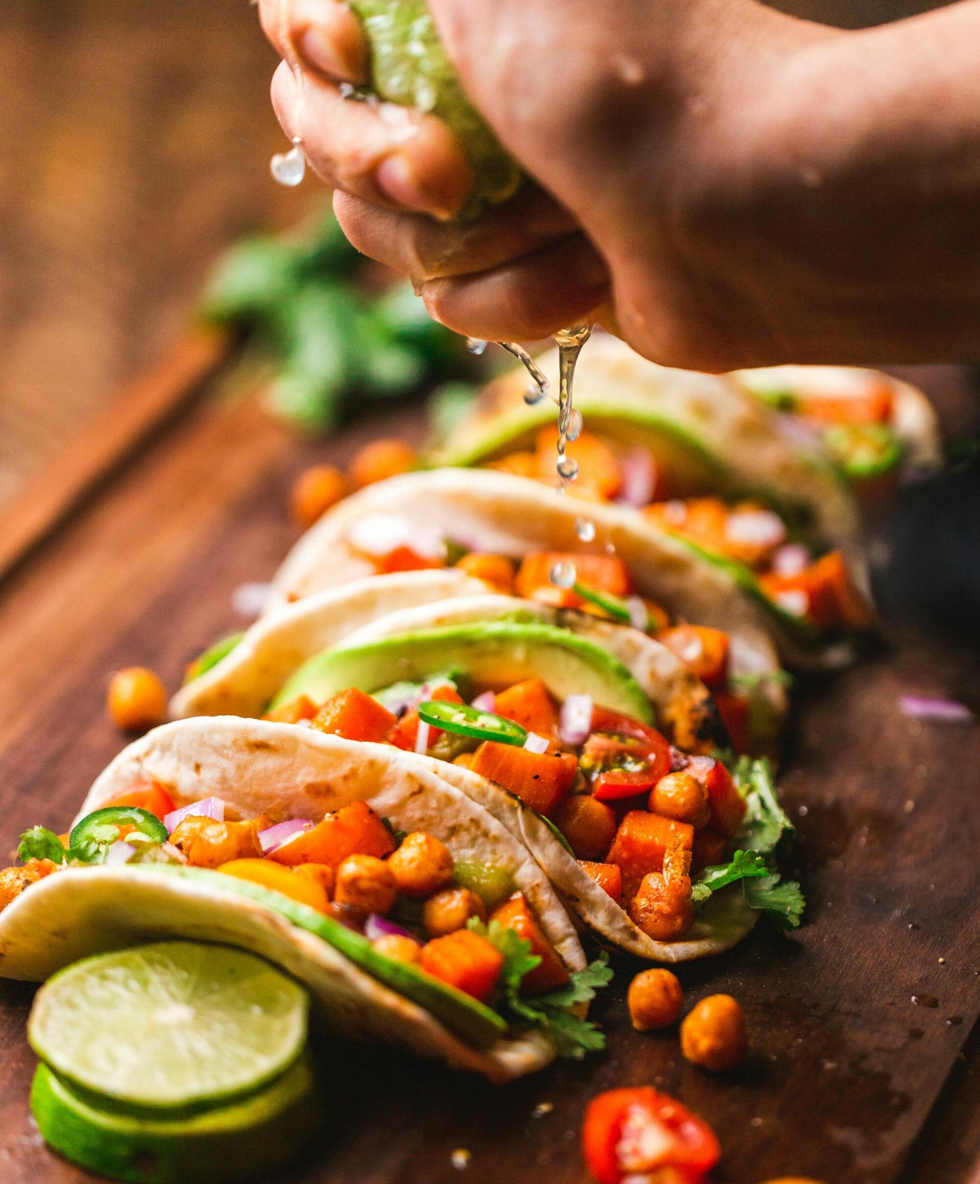 A hand squeezes lime over vegetable-filled tacos on a wooden board, garnished with fresh herbs.