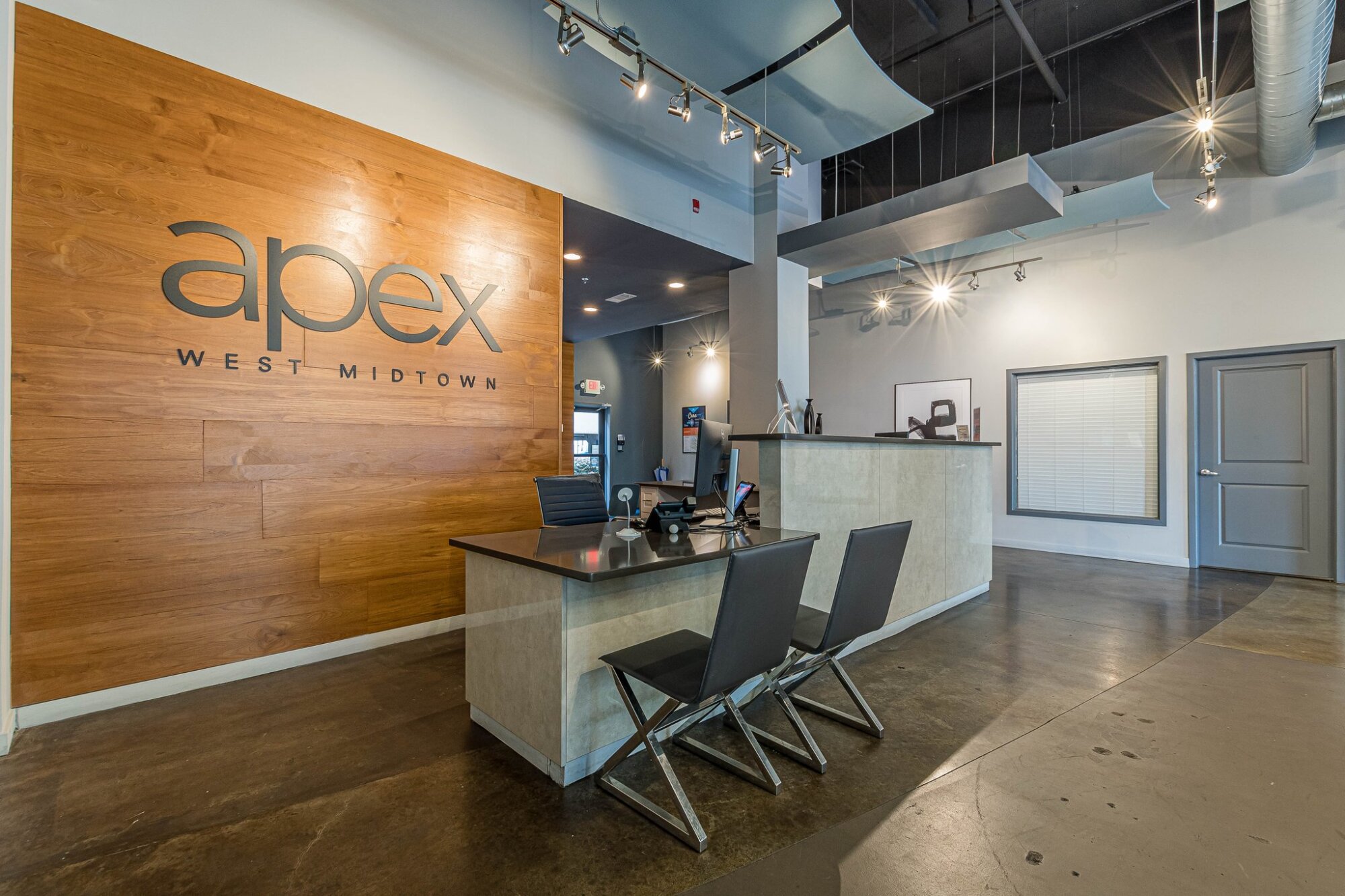 Modern apartment lobby at Apex West Midtown with gray sofas, a glass table, and "apex west midtown" sign on a wood accent wall.