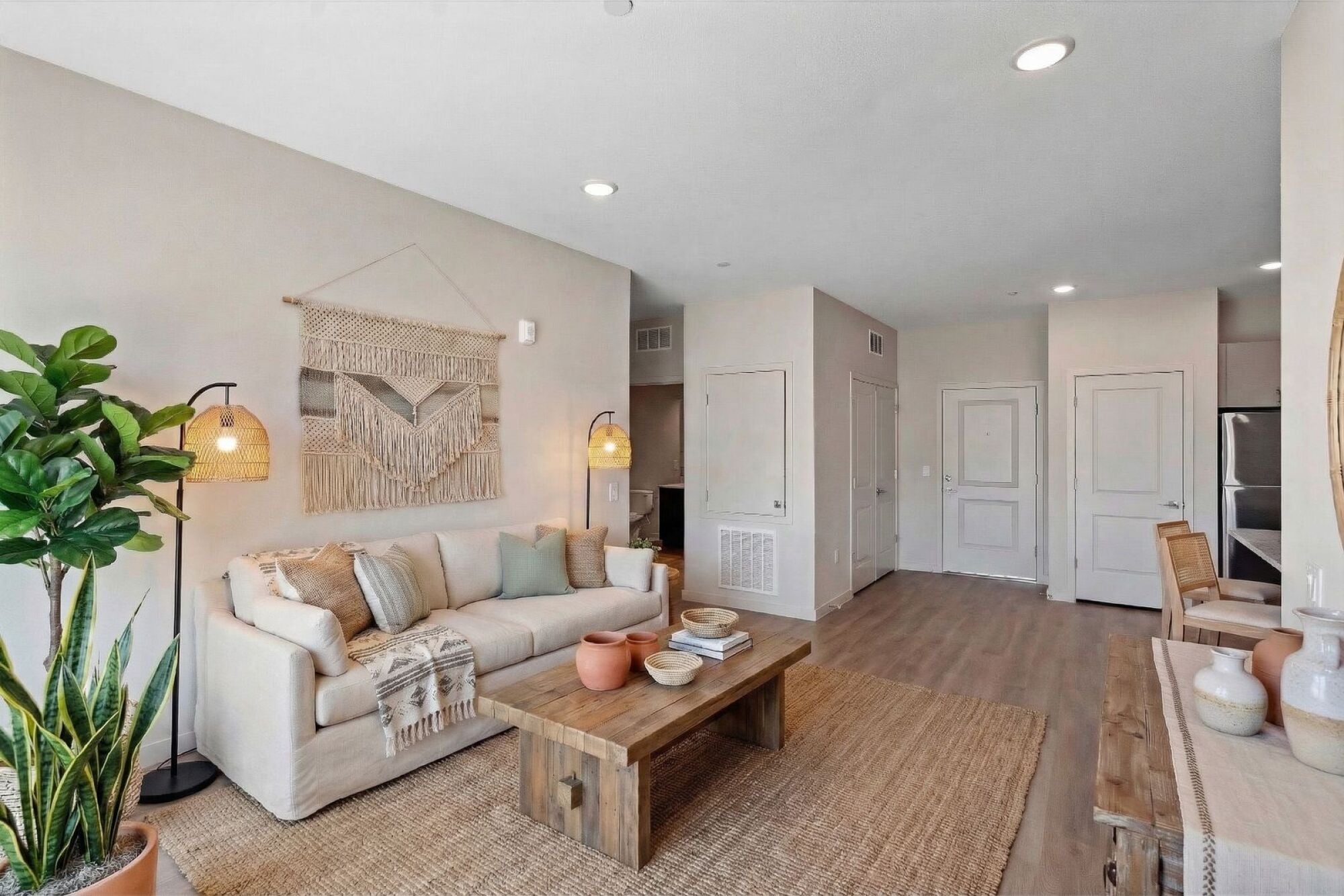 Neutral-toned living room with a beige sofa, wooden tables, plants, and woven decor on the wall.